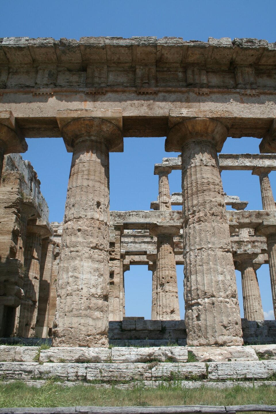 Fotografia nieznanego autora przedstawia widok na ruiny świątyni Posejdona w stylu doryckim. Budowla zbudowana jest z licznych kamiennych kolumn, które podtrzymywały dach budynku. Fotografia została wykonana w jasny i bezchmurny dzień. 