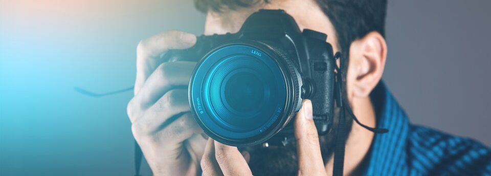 Ilustracja przedstawia fotografa w trakcie pracy. Mężczyzna widoczny jest od barków w górę. Ma czarną brodę oraz włosy. Ubrany jest w niebiesko‑granatową koszulę w drobną kratkę. Patrzy przez wizjer aparatu, jedną dłonią obejmując korpus oraz spust migawki, a drugą regulując obiektyw.