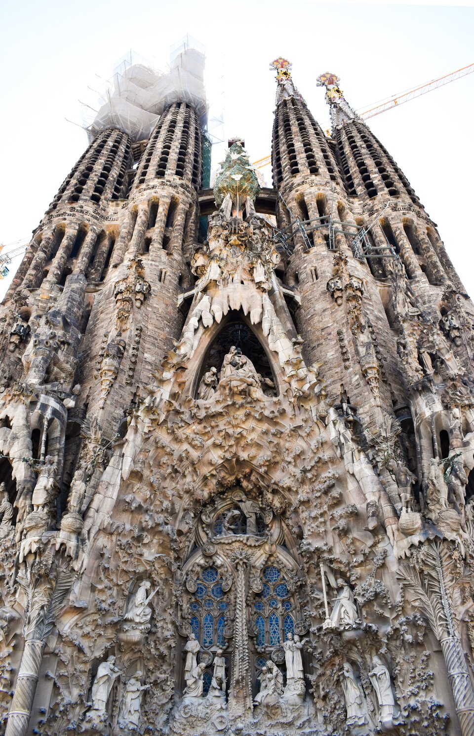 Ilustracja przedstawia Fasadę Narodzenia Pańskiego, kościół Sagrada Família, Barcelona, Hiszpania. Ukazuje niezwykle bogato rzeźbioną fasadę świątyni, pełną kamiennych figur, scen biblijnych i motywów roślinnych. Świątynia posiada strzeliste, ażurowe wieże,które zdobią liczne detale wyrzeźbione w jasnym kamieniu, tworzące bardzo złożoną, niemal koronkową strukturę. W dolnej części fasady znajdują się wejścia otoczone dekoracyjnymi reliefami, a wyżej - okna z witrażami. W górnych partiach dostrzegalne są rusztowania, świadczące o trwających pracach budowlanych.