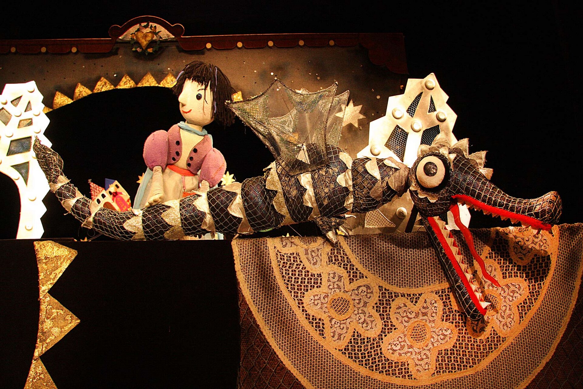 Ilustracja przedstawia kadr ze spektaklu teatralnego. Na fotografii widoczne są kukiełki. Z lewej strony kukiełka o brązowych, krótkich włosach z włóczki, w jasnoczerwonym kubraczku, patrzy w lewą stronę. Przed nią znajduje się długi złoto‑brązowy smok ze złotymi skrzydłami. Ma otwartą paszczę z czerwonym, długim językiem. Smok ma łuski zrobione ze złotego sznurka. W tle widać pieczarę smoka.