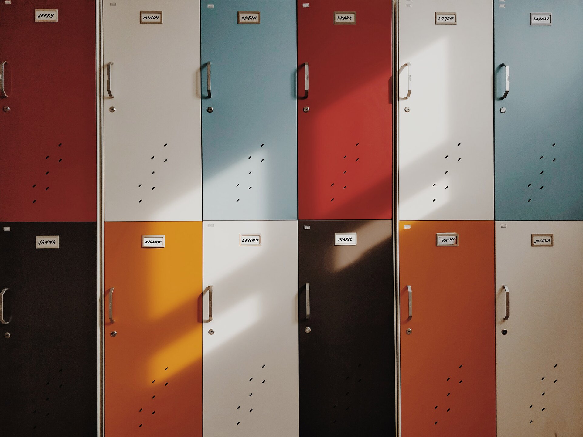 Zdjęcie przedstawia dwa rzędy drzwi od szafek szkolnych , w każdym rzędzie jest po sześć sztuk. Drzwi szafek są w różnych kolorach i znajdują się na nich karteczki z imionami. Każda z szafek posiada rączkę do otwierania a pod nią umieszczony zamek na kluczyk.