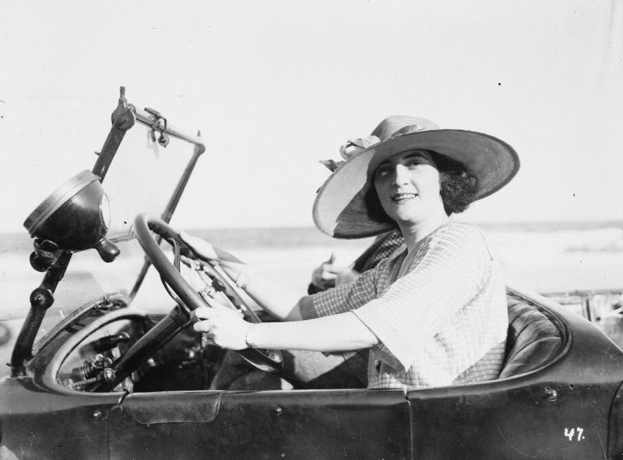 Zdjęcie przedstawia młodą kobietę, która prowadzi samochód. Ma na głowie kapelusz z szerokim rondem i uśmiecha się.