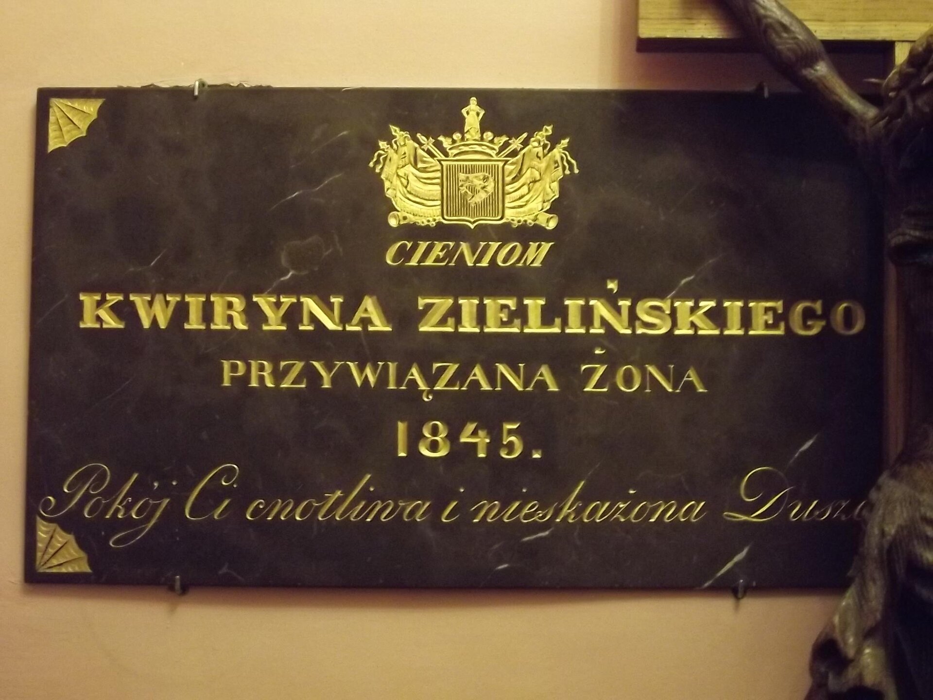 Zdjęcie przedstawia czarną tablicę pamiątkową, na której znajduje się napis. Na samej górze tablicy umieszczony został złoty herb, a poniżej znajduje się epitafium: "Cieniom. Kwiryna Zielińskiego, przywiązana żona. 1845. Pokój Ci cnotliwa i nieskażona Duszo". Tablica wisi na kremowej ścianie.