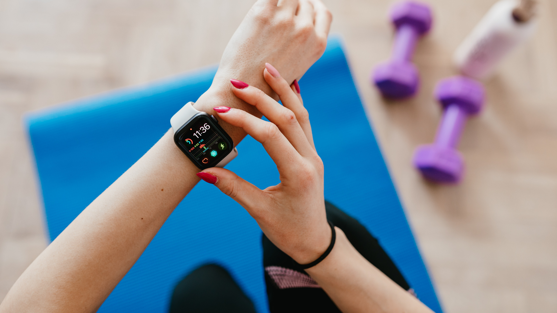 Zdjęcie wykonane z góry przedstawia zbliżenie na ręce kobiety wykonującej ćwiczenia na specjalnej macie do ćwiczeń. Na lewym ręku kobieta ma zegarek typu smartwatch. Prawą ręką kobieta naciska przycisk na zegarku. Na drugim planie zdjęcie jest lekko rozmyte. Widoczna jest mata do ćwiczeń. Obok maty na podłodze leżą dwa hantle i butelka z napojem.