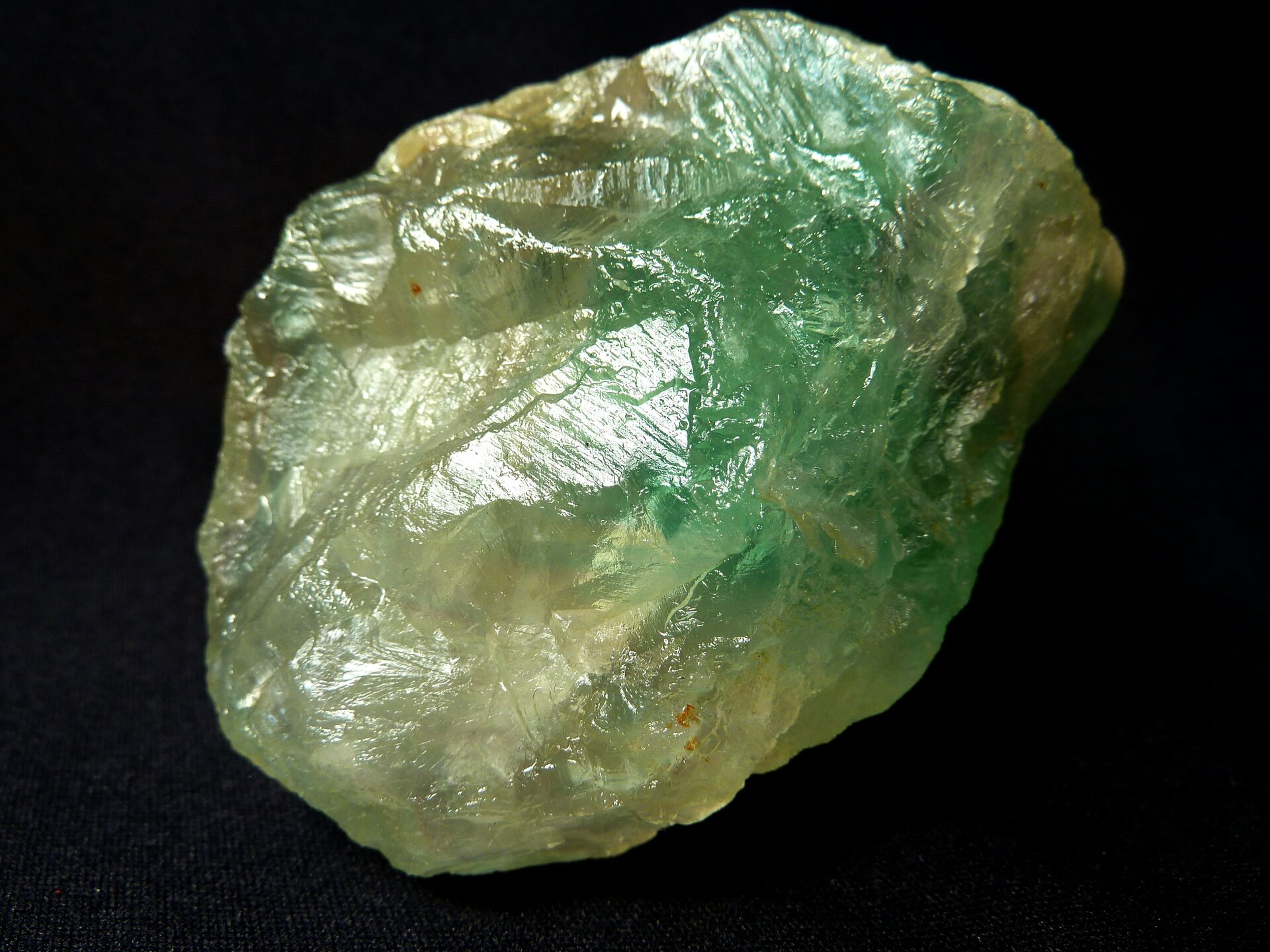 Ilustracja przedstawia fluoryt, kształtem przypomina kamień o śliskiej powierzchni, przypominającej szkło. Ma zielonkawy kolor. 