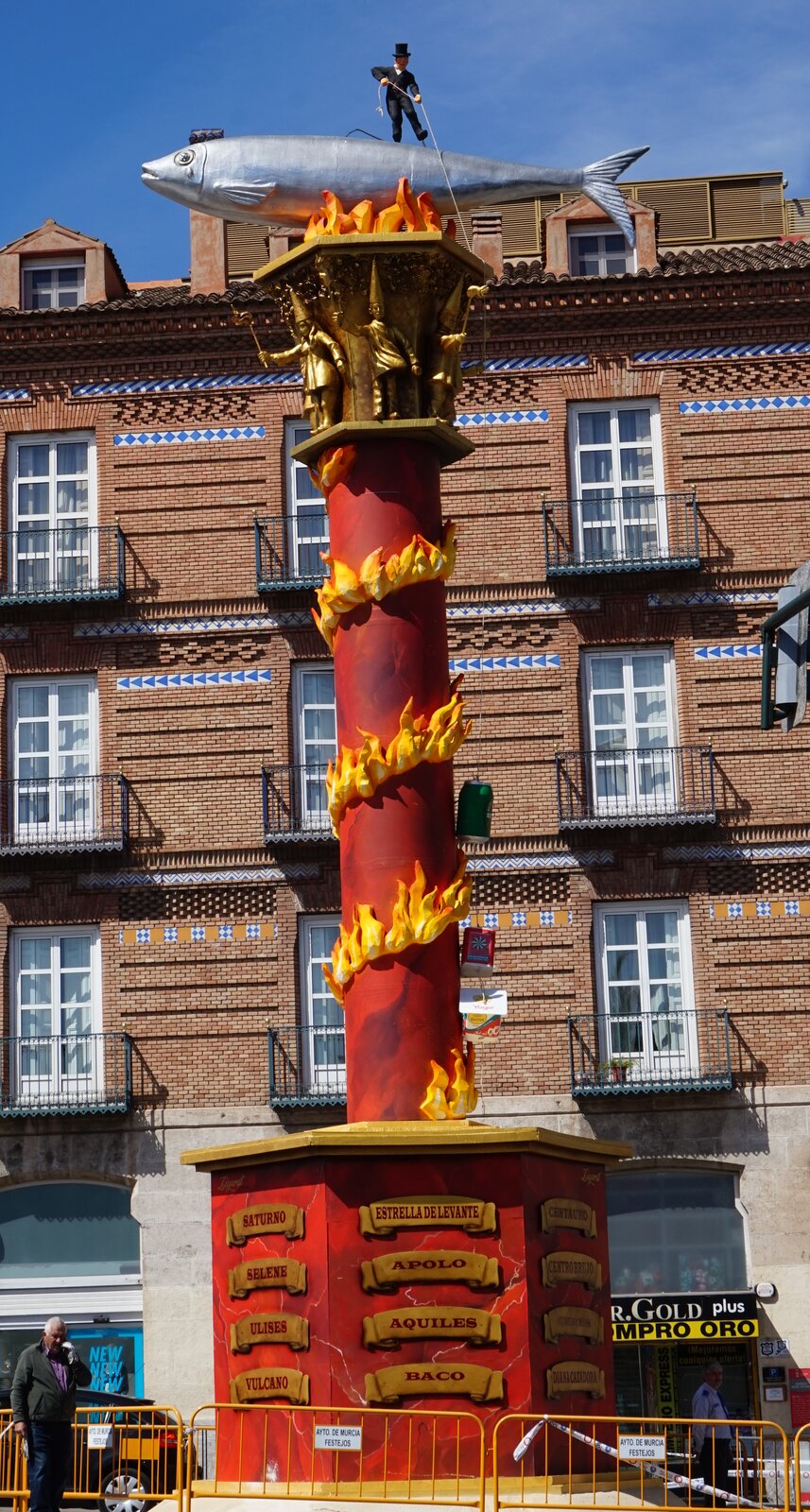 Zdjęcie przedstawia wysoką czerwoną wieżę, po której pnie się sztuczny płomień. Na szczycie znajduje się duża sardynka nad ogniem, a na jej grzbiecie posąg mężczyzny w czarnym cylindrze ciągnącego linę. W tle ceglany budynek.