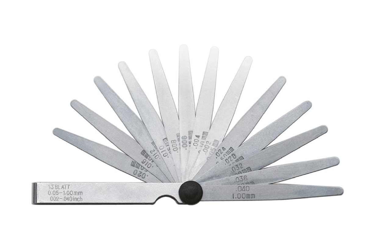 Ilustracja przedstawia urządzenie. To szczelinomierz. Jest on zbudowany z szeregu podłużnych płytek o różnej grubości, które osadzone są we wspólnej obudowie i spięte stalową osią.