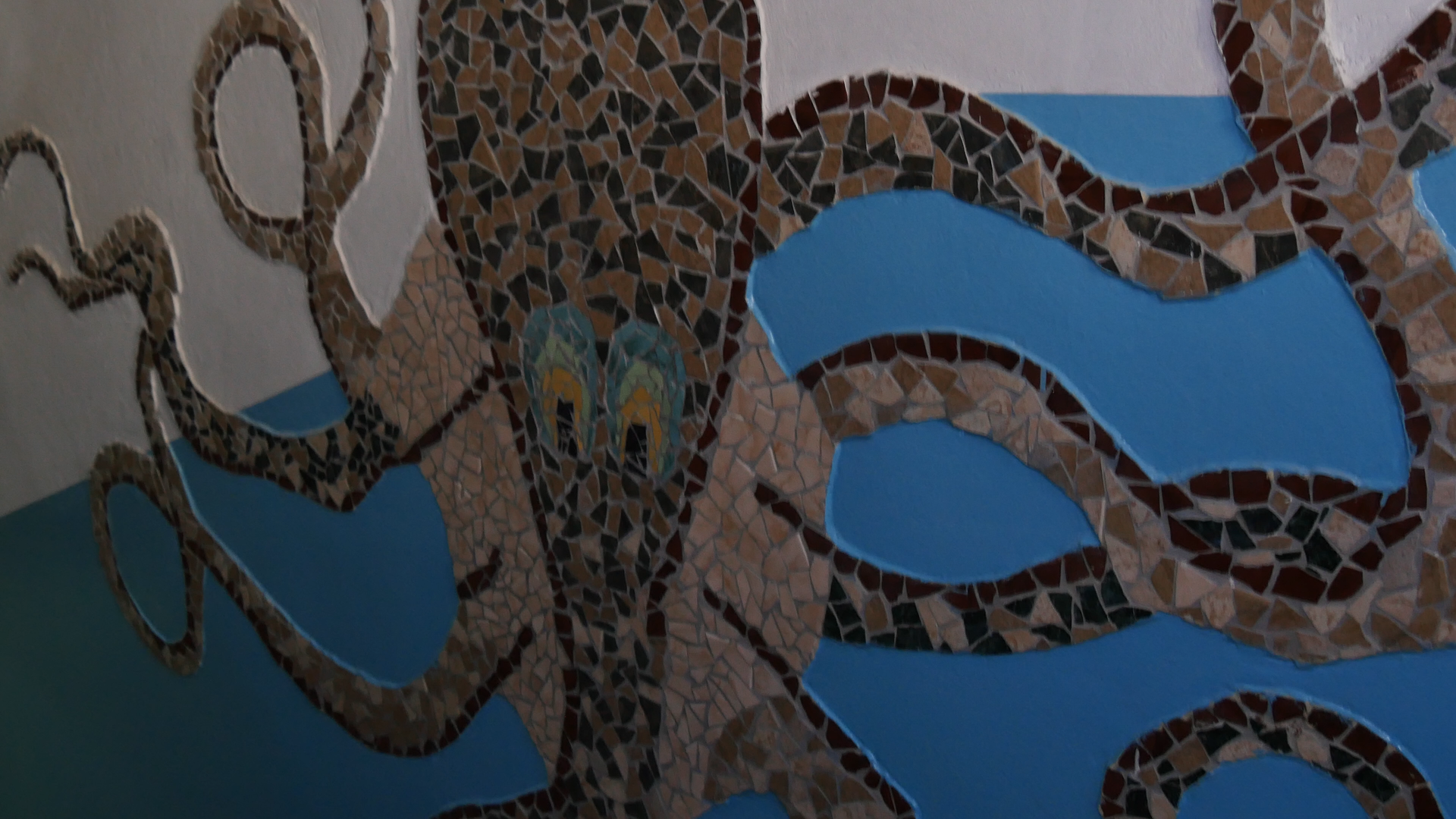 Zdjęcie przedstawia model detalu architektonicznego wykonany z ceramiki. Model składa się z drobnych ceramicznych płytek w różnych kolorach i przedstawia ośmiornicę. Jest wkomponowany w biało‑niebieską ścianę.