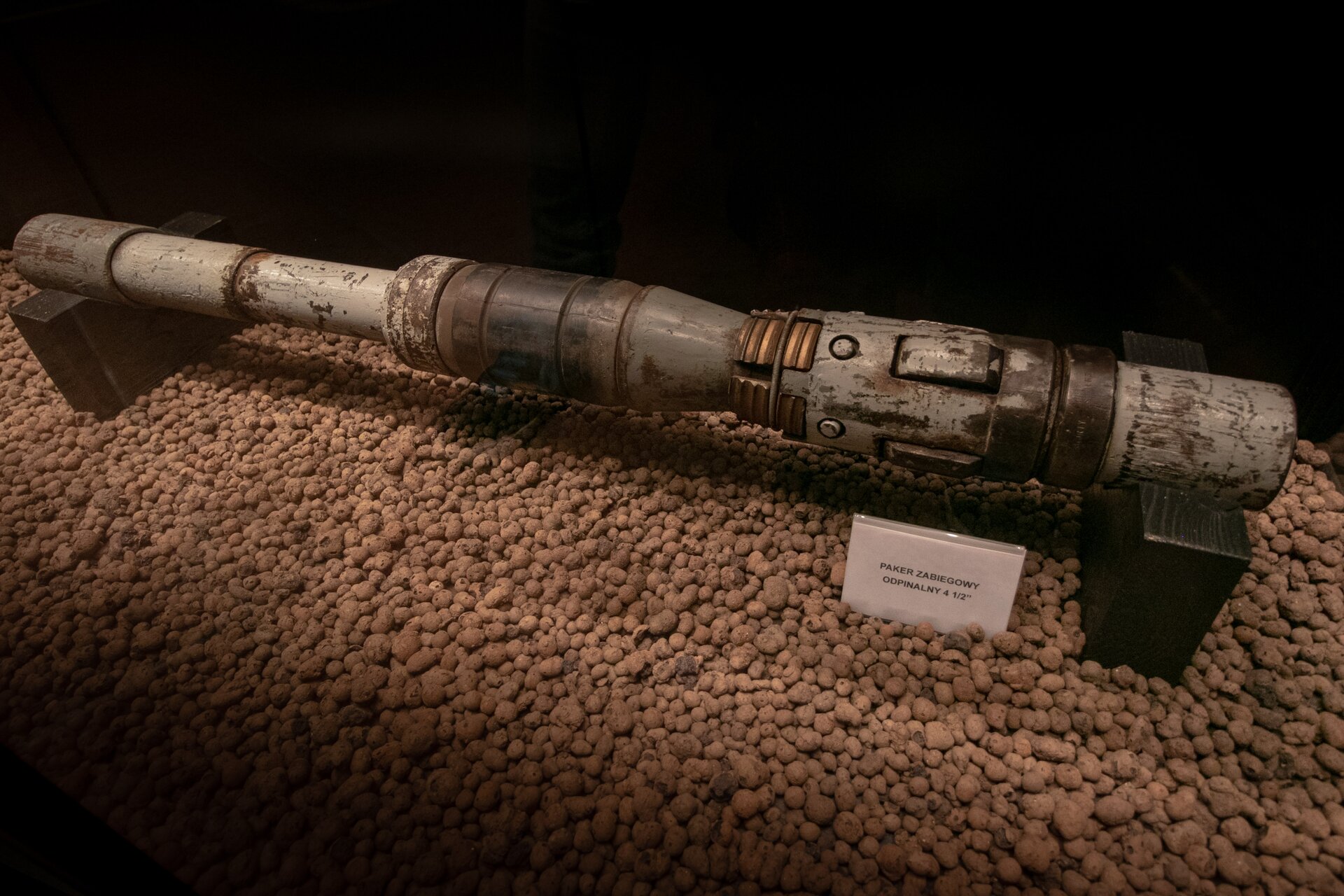 Na zdjęciu widoczny jest szary paker zabiegowy. Paker jest to narzędzie wgłębne stosowane w otworach wiertniczych i odwiertach wydobywczych. Umożliwiające uszczelnienie przestrzenia pierścieniowej między przewodem wiertniczym a ścianą otworu lub rurami okładzinowymi, albo między rurami wydobywczymi a rurami okładzinowymi w odwiercie wydobywczym, w celu uniemożliwienia przepływu pionowego płynu złożowego przez uszczelnioną przestrzeń pierścieniową między rurami.Widoczny na zdjęciu paker jest urządzeniem o kształcie podłużnego cylindra którego można podzielić na cztery części. Pierwszy element po prawej stronie jest o kształcie szarego metalowego cylindra. Drugi element jest o kształcie walca którego powierzchnia boczna jest wypukła Element ten jest najszerszy w porównaniu z całym przekrojem urządzenia. W dolnej części elementu widać równomiernie rozłożone po obwodzie okręgi. Trzeci element jest o kształcie walca którego krawędzie przy prawej podstawie są nieznacznie ścięte. Czwarty element jest o kształcie teleskopowego walca który zbudowany jest z trzech mniejszych elementów. Każdy z nich jest o nieznacznie większej średnicy.Paker posadowiony jest na dwóch drewnianych klockach. Na środku klocków wycięty jest otwór w kształcie koryta. Klocki umieszczone są na obu krańcach urządzenia. Drewniane klocki są posadowione na gruncie ma którym wysypane są brązowe kamienie. Przed pakerem znajduje się biała tabliczka z czarnym opisem. Tabliczka umieszczona jest po prawej stronie pakera.