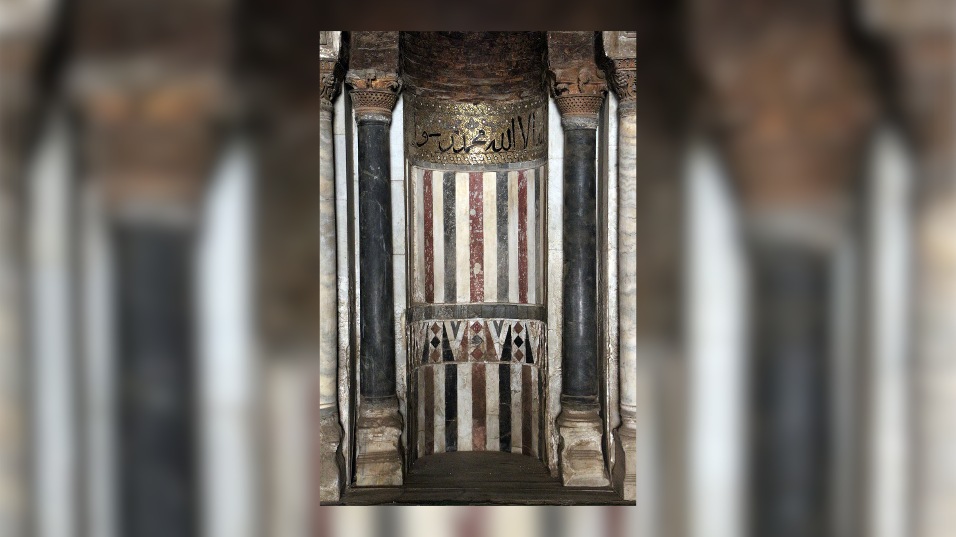 Na zdjęcie znajduje się Mihrab. Jest to wnęka w ścianie, po obu jej stronach znajdują się ozdobne ciemne kolumny. wnęka jest ozdobiona malowidłami figur geometrycznych, a w jej górnej części na złotym tle znajdują się czarne napisy zapisane pismem arabskim. 