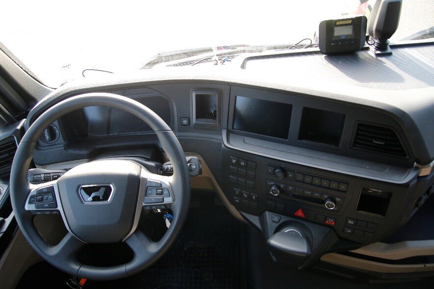 Ilustracja przedstawia deskę rozdzielczą z widoku kierowcy, widoczna jest kierownica za kierownicą wyświetlacz układu sterowania samochodu. po prawej stronie znajduję się mniejszy ekran. poniżej wiele przycisków na przykład przyciski do obsługi radia lub kontrolowania ogrzewania w aucie. Pod przyciskami znajduję się gałka do zmienia biegów.