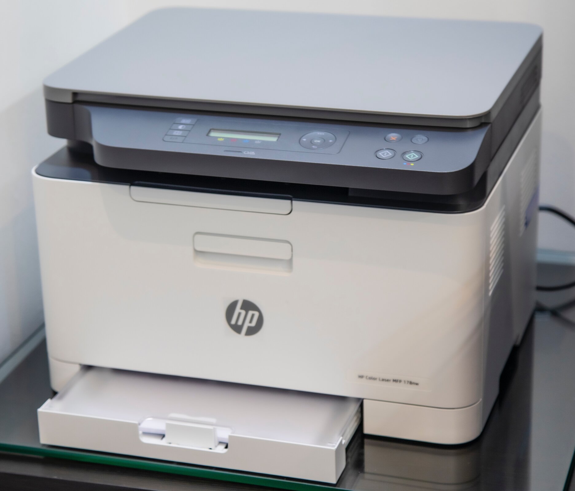 Zdjęcie przedstawia wielofunkcyjne urządzenie łączące w sobie funkcje drukarki i skanera. Widoczna jest prostopadłościenna szaro‑biała obudowa urządzenia, w dolnej części widoczna jest lekko wysunięta szuflada, w której widoczny jest włożony papier. W górnej części urządzenia widoczna jest szara klapa, z panelem sterującym z przyciskami i diodami. Na przodzie w środkowej części obudowy, widoczny jest płaski uchwyt. Urządzenie stoi na blacie, a w tle po prawej stronie widoczne są czarne kable.