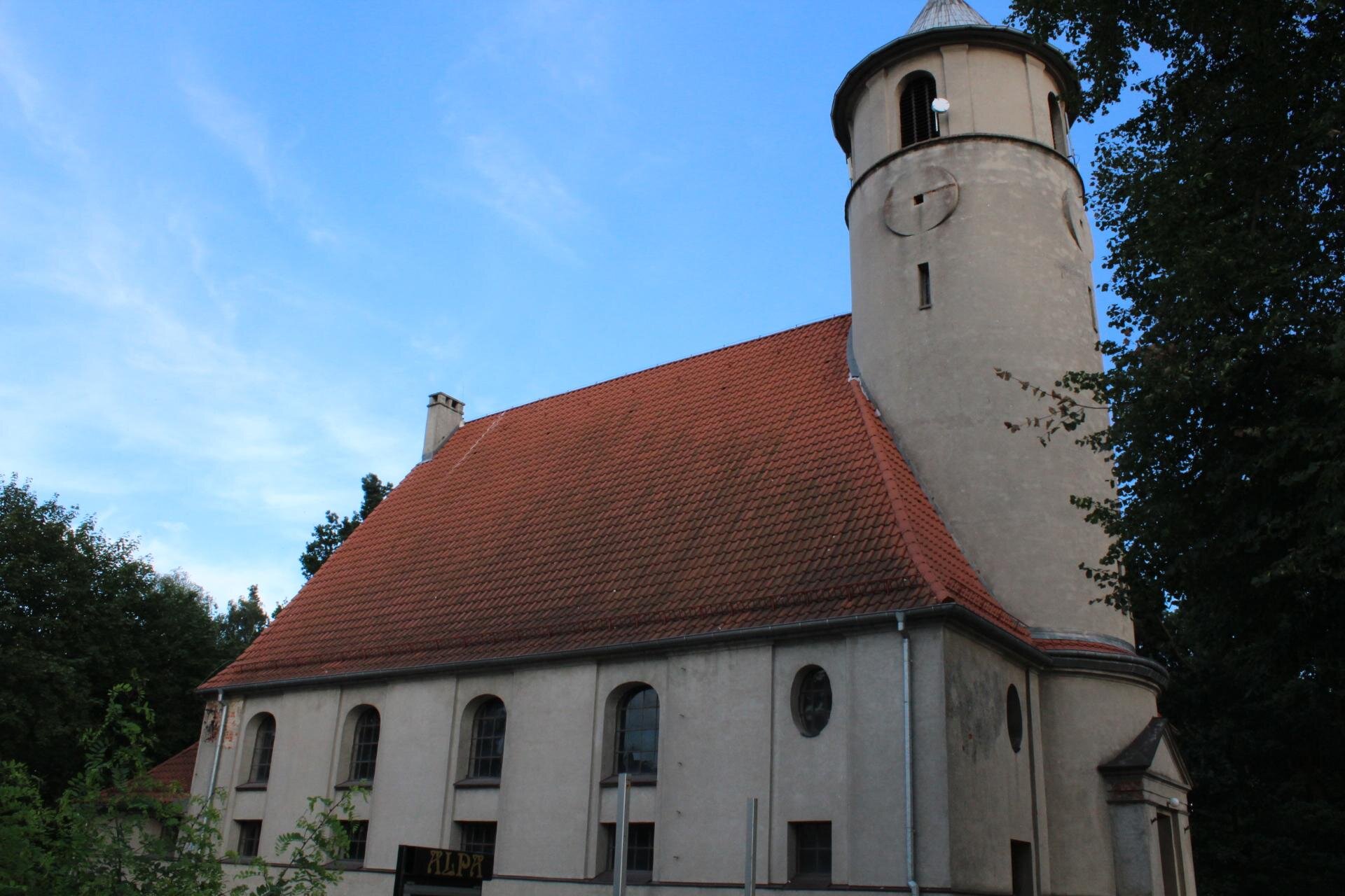 Zdjęcie przedstawia kremowy kościół z pomarańczowym dachem. Okna są zaokrąglone u góry. Z przodu kościoła stoi wysoka wieża. Wokół budynku, z prawej i lewej strony zdjęcia, widoczne są zielone fragmenty drzew. Niebo jest błękitne. Zdjęcie zostało wykonane w świetle dziennym.
