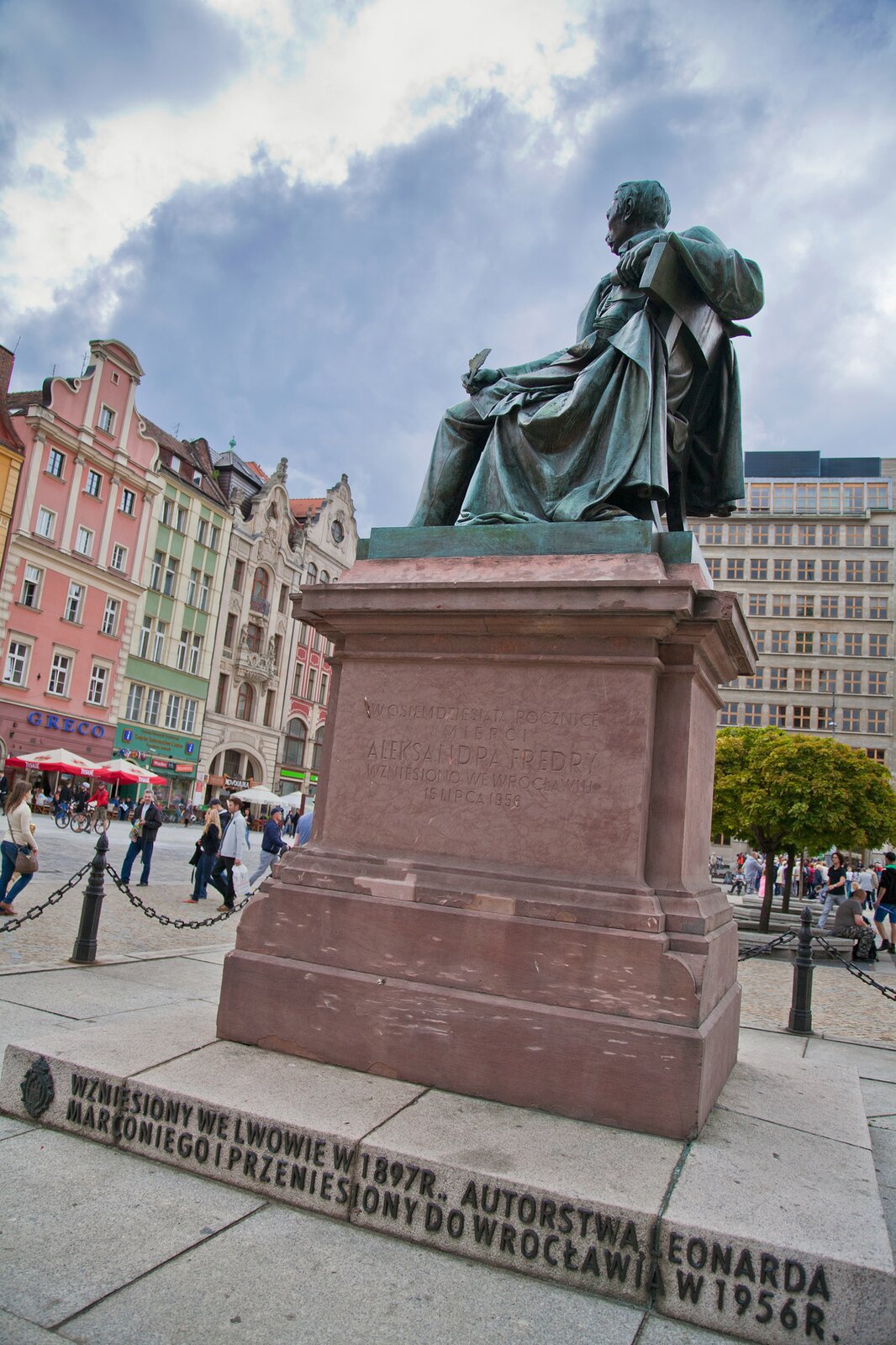 Zdjęcie przedstawia prawy bok pomnika siedzącego na krześle pisarza, który w jednej dłoni trzyma pióro, a w drugiej rolkę papieru. Rzeźba ustawiona jest na wysokim, marmurowym cokole. Na prawym boku cokołu wygrawerowano: W osiemdziesiątą rocznicę śmierci Aleksandra Fredry wzniesiono we Wrocławiu 15 lipca 1956. Poniżej, na stopniu pod pomnikiem znajduje się napis: Wzniesiony we Lwowie w 1897 r., autorstwa Leonarda Marconiego i przeniesiony do Wrocławia w 1956 r. W tle widać kamienice.
