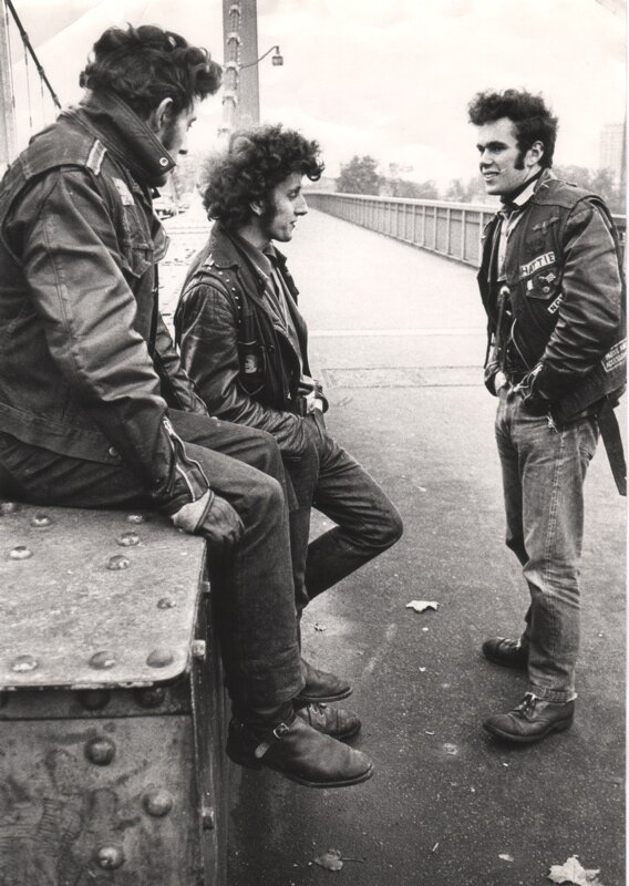 Zdjęcie przedstawia trzech młodych mężczyzn. Znajdują się na moście. Są ubrani w skórzane kurtki, mają założone rękawiczki oraz ciemne, ufryzowane włosy.