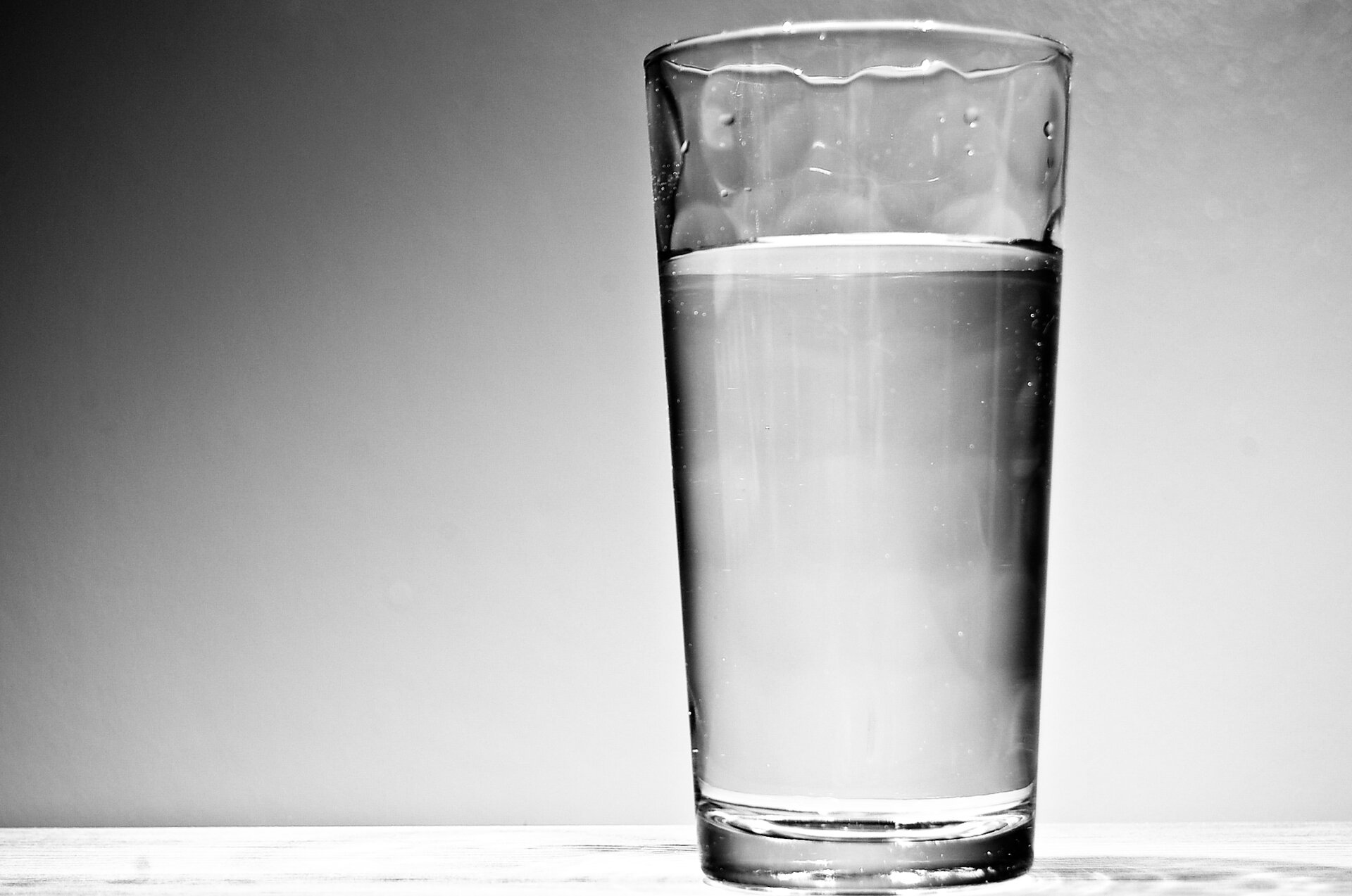 Grafika przedstawia zdjęcie, na którym znajduje się przezroczysta szklanka wypełniona wodą. Szklanka przedstawiona jest na szarym tle.