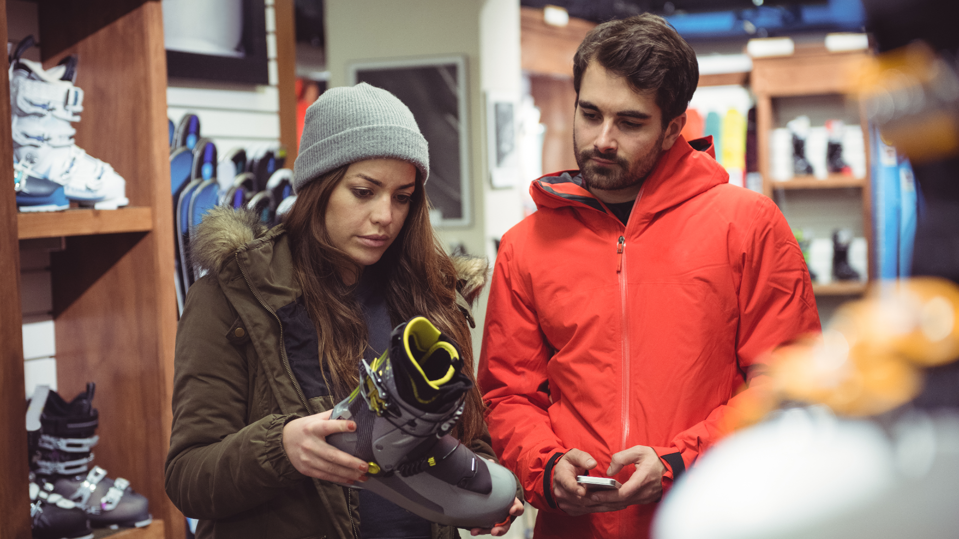 Zdjęcie przedstawia kobietę i mężczyznę w sklepie sportowym. Kobieta znajduje się po lewej stronie i trzyma w ręku but narciarski. Obok niej po prawej stoi mężczyzna.