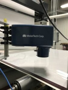 Zdjęcie przedstawia czujnik. Widoczne jest urządzenie złożone z prostopadłościennego elementu, na końcu którego od dołu widoczny jest cylindryczny element. Element ten znajduje się nad biały blatem. Prostopadłościenna część urządzenia przymocowana jest czterema zaczepami do pionowej metalowej rurki znajdującej się na brzegu blatu.