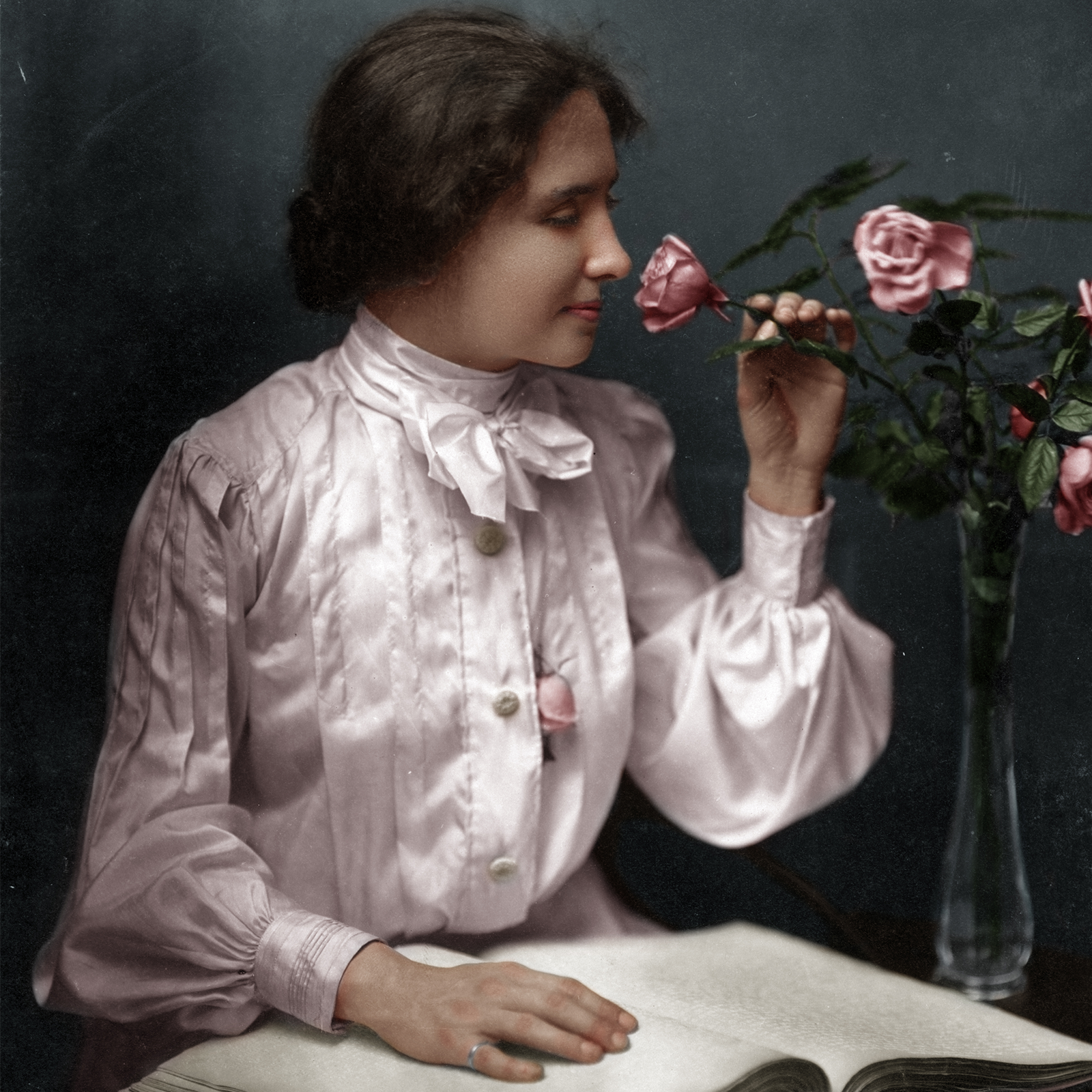 Zdjęcie przedstawia młodą kobietę siedzącą przy stole i wąchającą różę, która stoi w wazonie. Jej prawa dłoń położona jest na otwartej książce. 