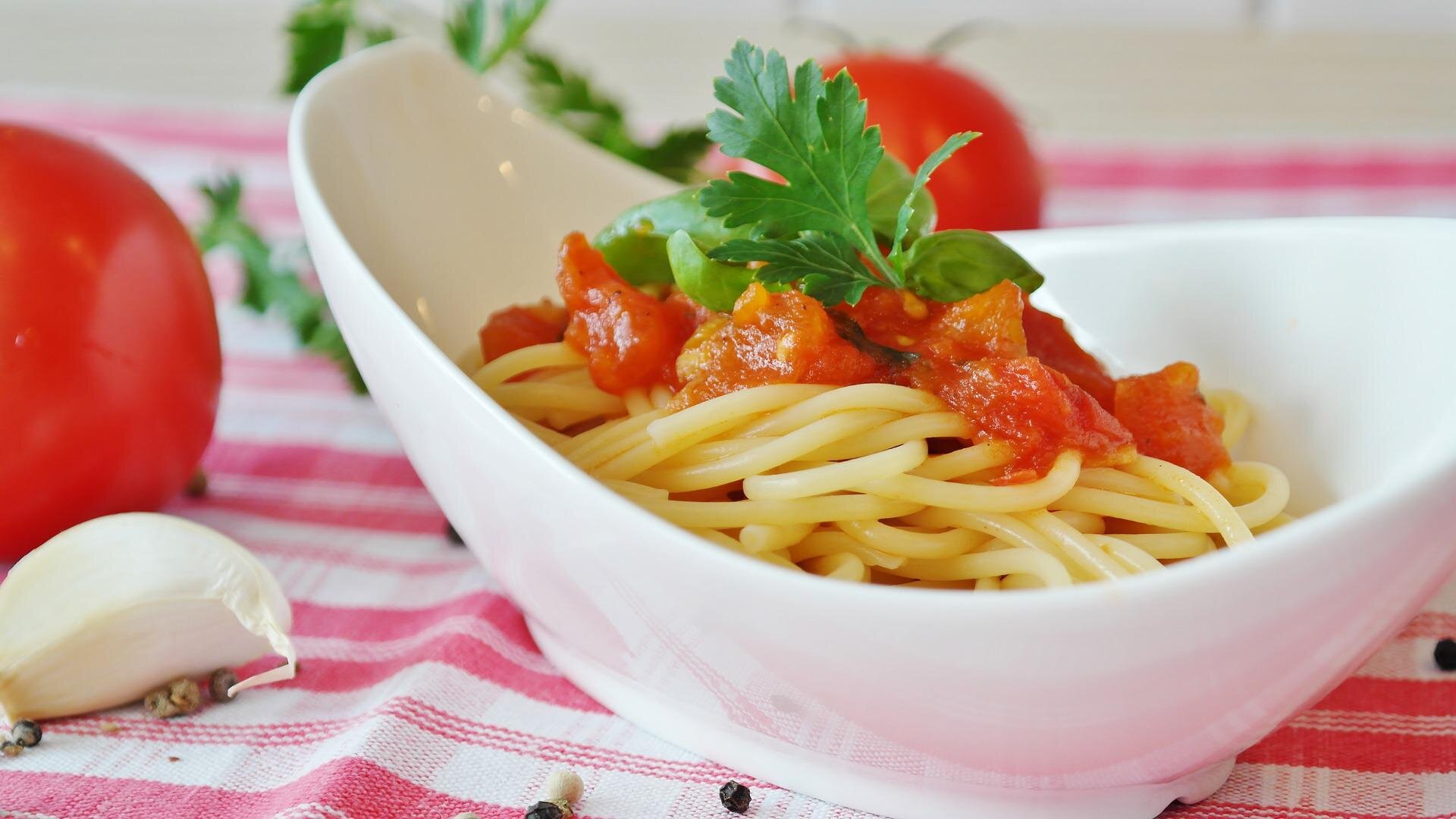 Zdjęcie przedstawia miseczkę, w której znajduje się makaron spaghetti polany na wierzchu sosem pomidorowym i przyozdobiony liśćmi pietruszki. Obok miseczki leżą ząbek czosnku, pomidory i ziarna pieprzu. 