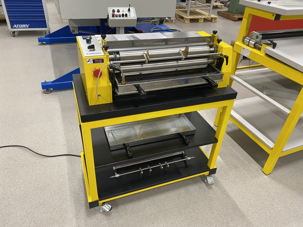 Na zdjęciu przedstawiono kaszerownicę. Jest to urządzenie osadzone na stabilnej metalowej ramie, wyposażone w system wałków oraz panel sterujący.