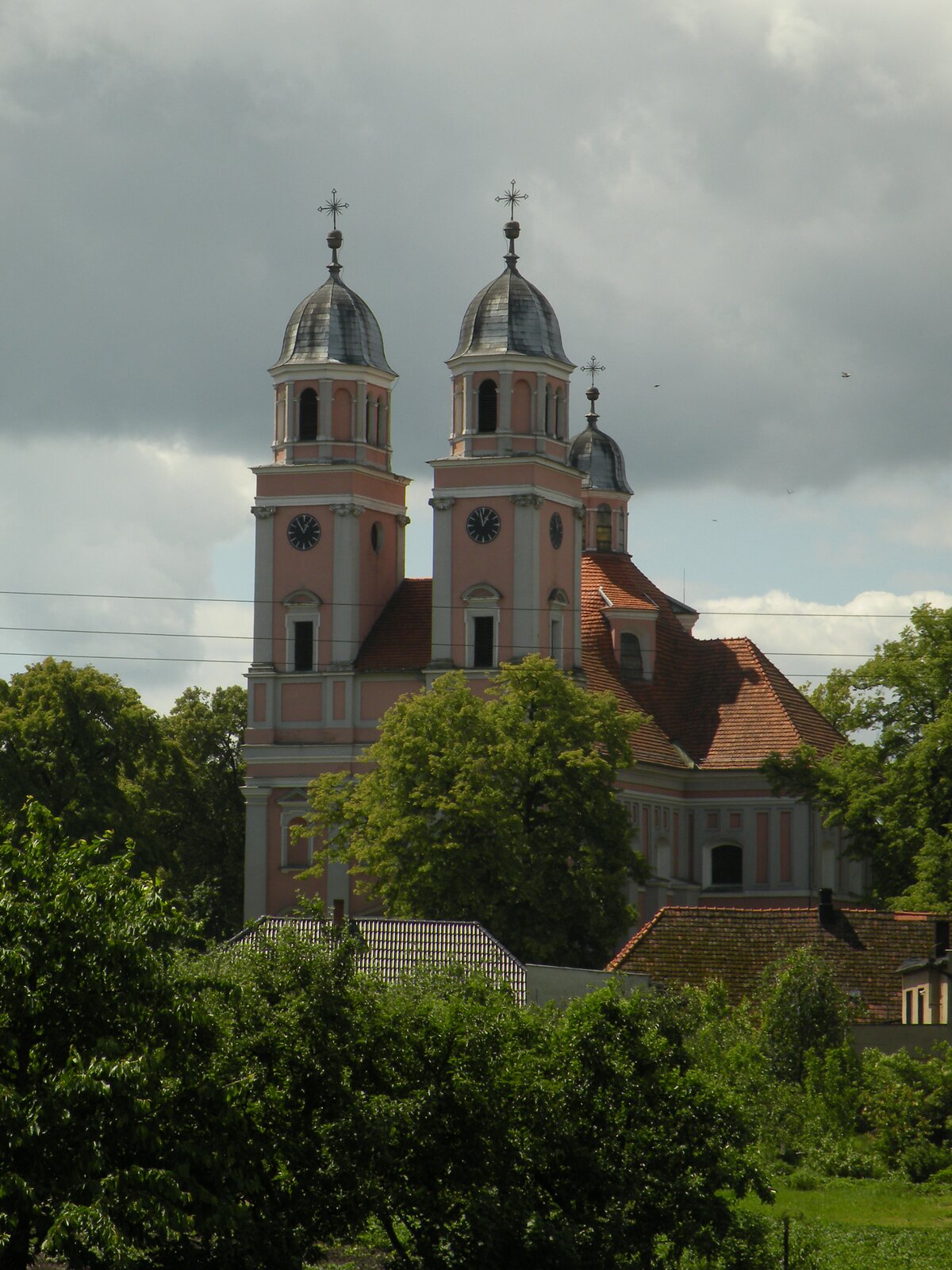 Zdjęcie przedstawia widok współczesny na kościół w Sierakowie. Wejście do wnętrza świątyni i fragment fasady są zasłonięte przez koronę drzewa. Widoczne są za to dwie wieżyczki od strony kruchty świątyni. Ich wystrój zewnętrzny opiera się na wątkach klasycystycznych i barokowych. Wieże mają podstawę na równi z początkiem wielospadowego dachu złożonego z dachówek. Są o podstawie kwadratu. W górnych partiach na fasadzie widoczne dwa zegary tarczowe, na wieży po prawej stronie widać zegar na bocznej ścianie. W szczytowej partii wieże zwężają się do formy wieloboków, nad którymi górują kopuły osłaniające część dzwonną. Na szczytach każdej z kopuł krzyże łacińskie. W głębi, za wieżami, widoczna sygnaturka o podobnym kształcie. Pod nią dach wielospadowy, z wykuszem w którym wbudowano otwór okienny. Boczne elementy kościoła, w tym skrzydło na prawo od nawy głównej również utrzymane w porządku klasycystycznym, poprzez wyznaczenie linii pilastrów. Gzyms pod dachem na całej długości fasady skrzydła, widoczny także fragment pod wieżą z lewej strony. Na pierwszym planie wielospadowe dachy dwóch domostw, częściowo zasłonięte przez drzewa i krzewy.