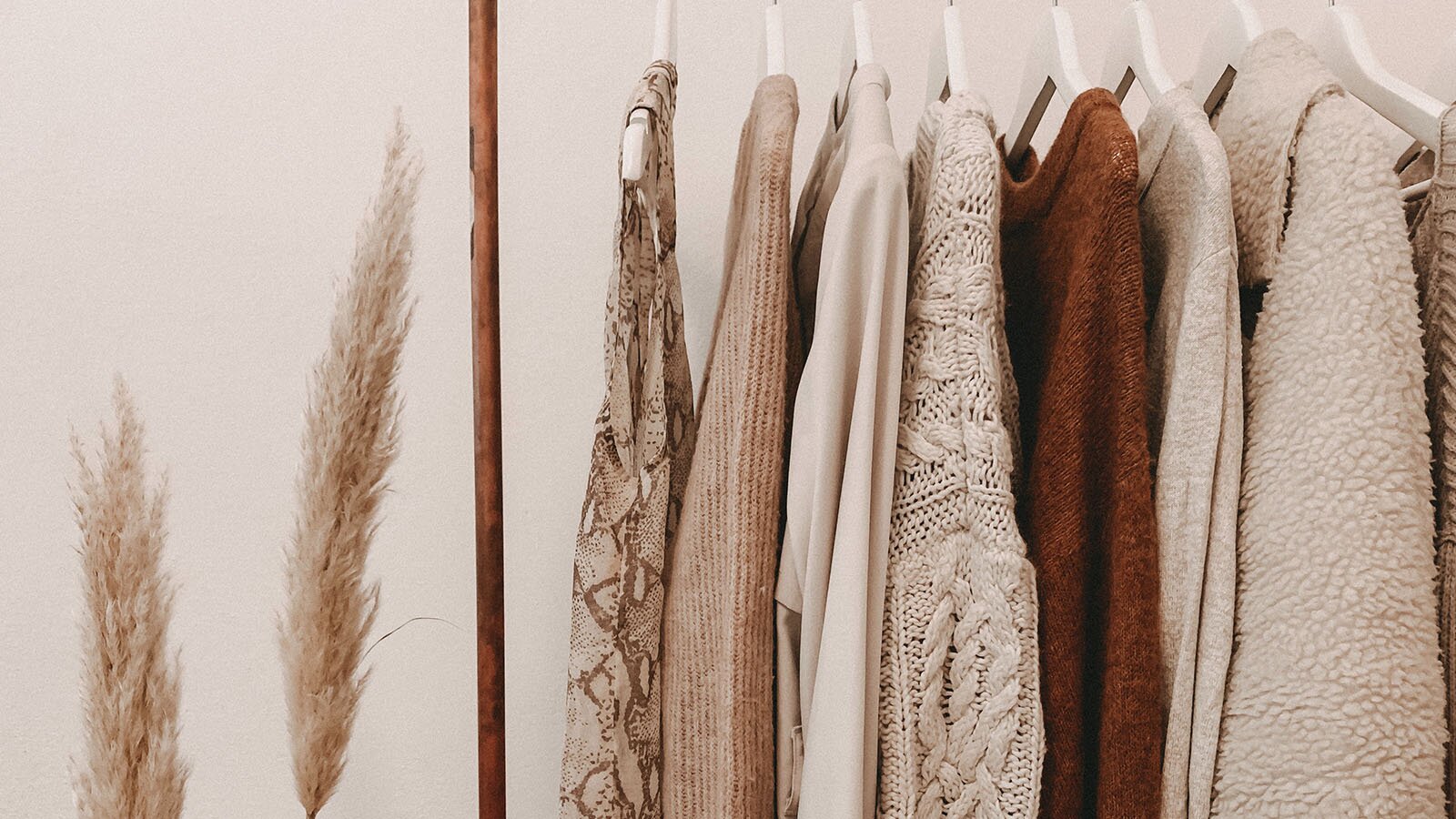 Zdjęcie przedstawia powieszone na wieszakach ubrania. Widoczny jest sweter, płaszcz i bluzki. Wszystko jest w stonowanych kolorach beżu, brązu i bieli. 
