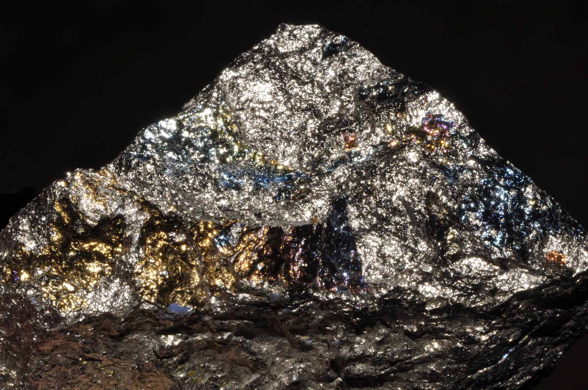 Zdjęcie prezentuje kawałek minerału. Ma zwartą budowę, połyskuje w kolorach srebrnym i zlotym oraz gdzieniegdzie niebieskim.