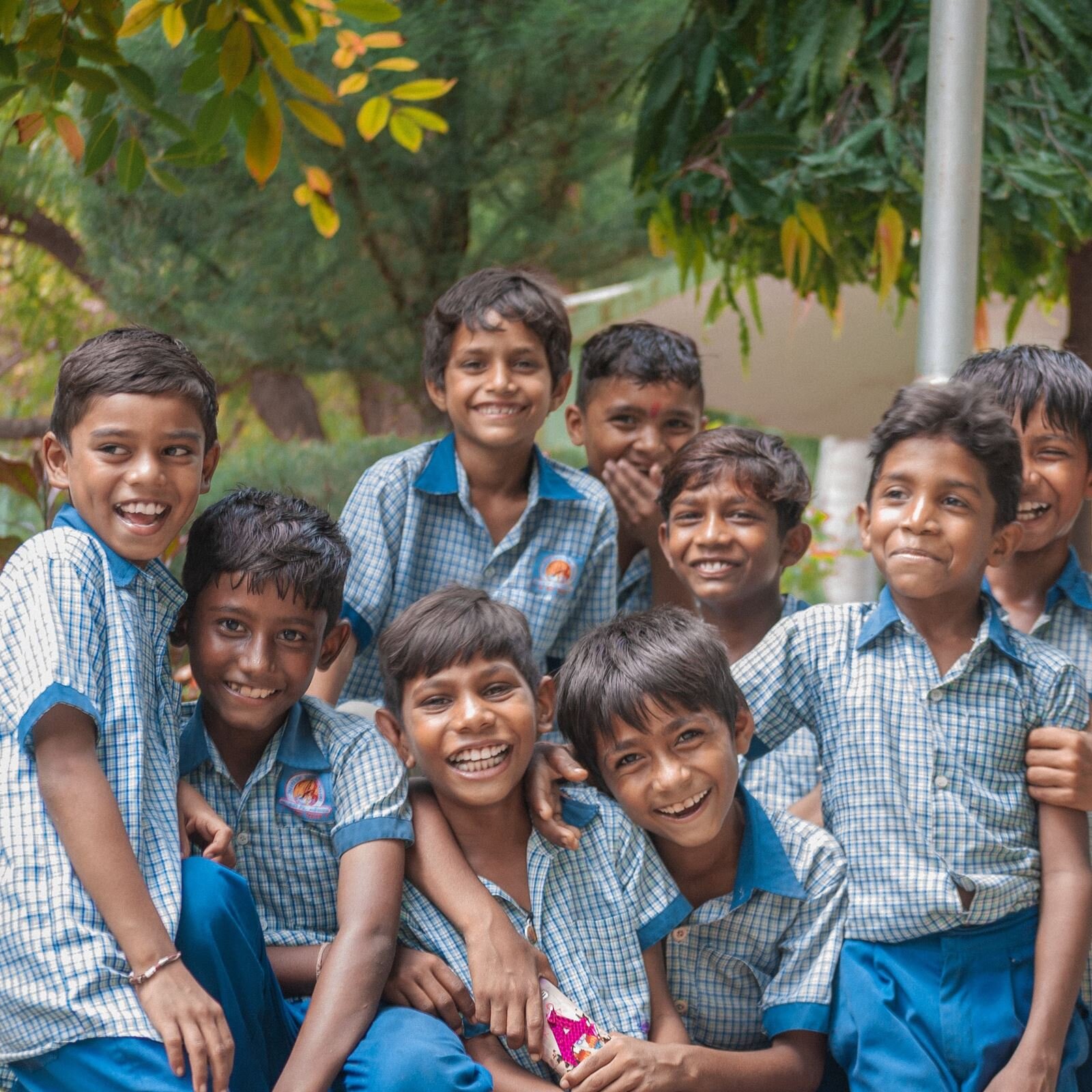 Zdjęcie przedstawia grupę 9 uśmiechniętych, ciemnoskórych chłopców, opierających się o siebie. Wszyscy ubrani są w jednakowe stroje. Koszule na krótki rękaw są w niebiesko-białą kratę. Kołnierzyk u koszuli jest niebieski.