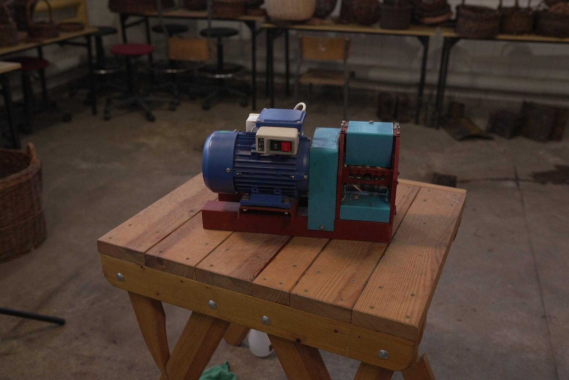 Zdjęcie przedstawia taśmiarkę elektryczną stojąca na drewnianym stole w pracowni koszykarskiej. Urządzenie składa się z silnika z włącznikiem i taśmiarki. 
