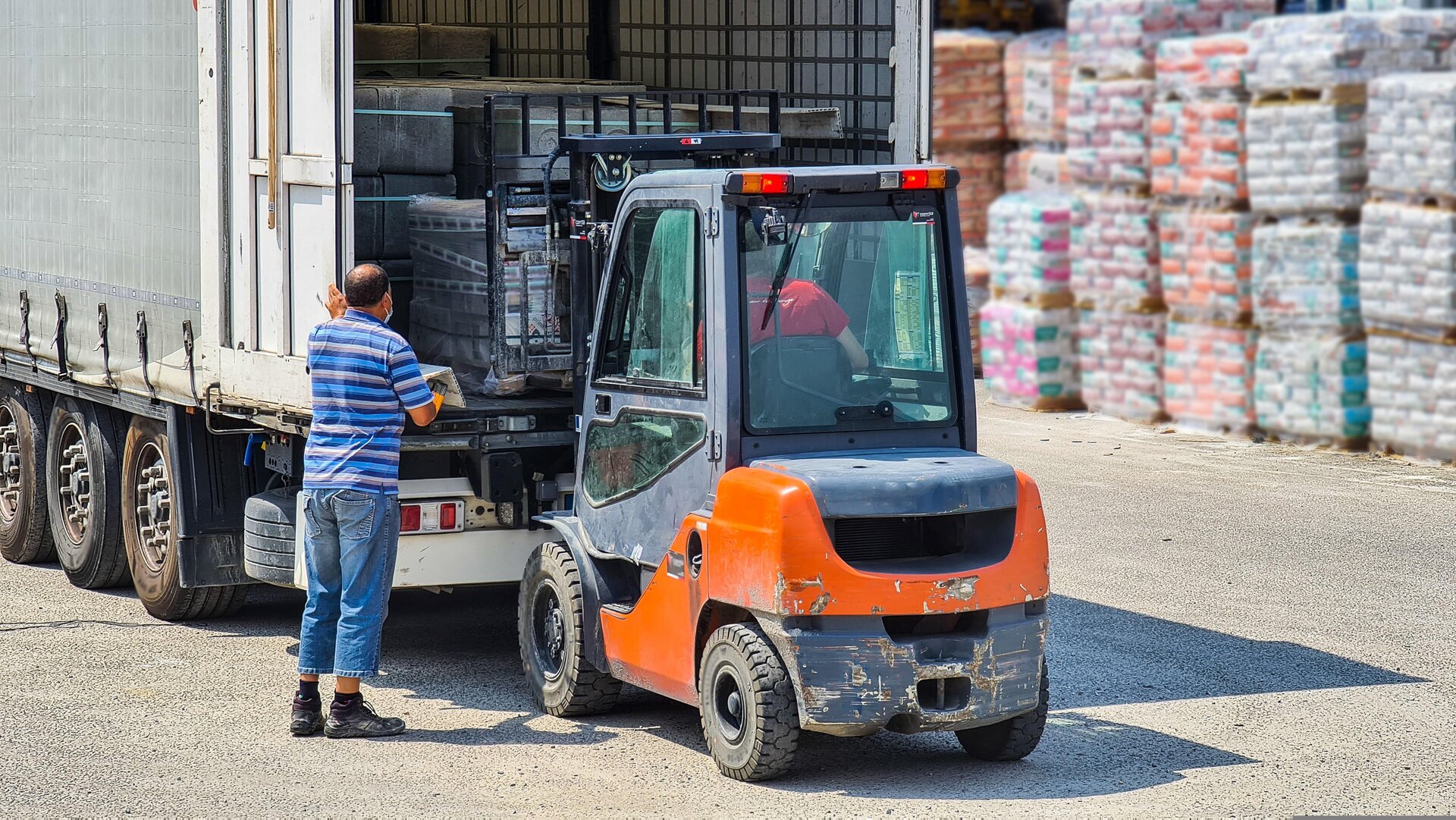 Zdjęcie przedstawia operację rozładunku. Paletowa jednostka ładunkowa wyciągana jest z naczepy za pomocą wózka widłowego kierowanego przez operatora. Proces ten nadzoruje kierowca, który stoi przy naczepie.