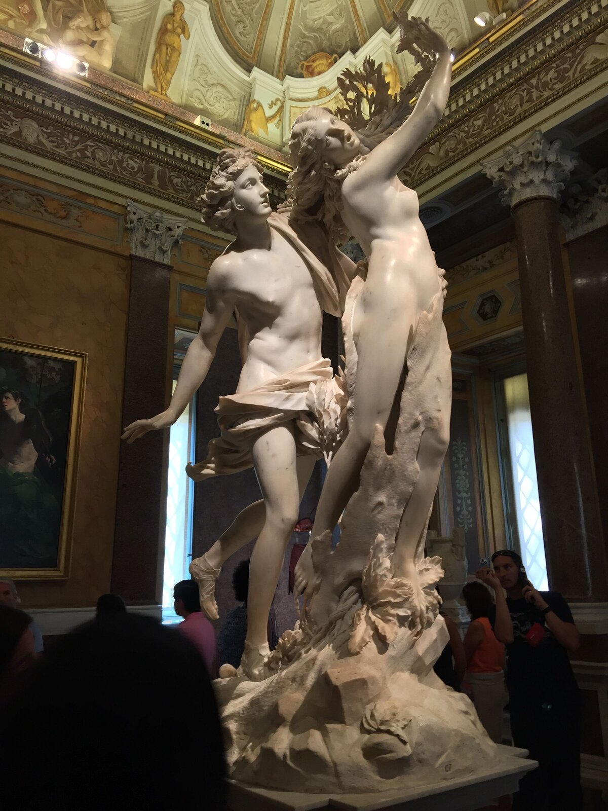 Ilustracja przedstawia rzeźbę „Apollo i Dafne” autorstwa Gianlorenzo Berniniego. Rzeźba ukazuje nagą kobietę oraz mężczyznę. Postać kobiety ma prawą rękę uniesioną do góry. Głowa jej jest przechylona w prawo, a długie włosy są rozwiane jakby przez wiatr. Postać ma przerażony wyraz twarzy. Mężczyzna stojący za kobietę, lewą ręką trzyma na jej biodrze. Postać również ma dłuższe, rozwiane włosy. Mężczyzna ma osłonięte krocze warstwą materiału. Jego prawa ręka wygięta jest do tyłu, a lewa stopa uniesiona do góry.