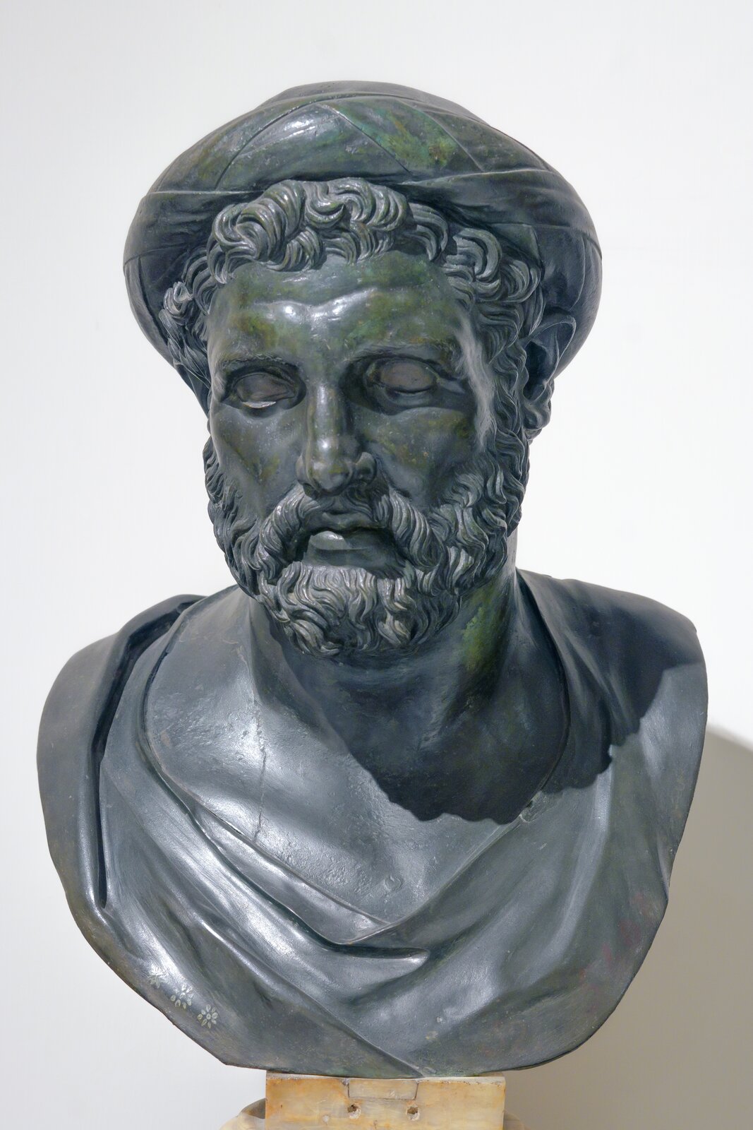 Zdjęcie przedstawia popiersie Pitagorasa, wykonane z ciemnego kamienia. Mężczyzna ma średniej długości kręconą brodę oraz wąsy. Spod okrągłego nakrycia głowy wystają kręcone włosy. Na jego czole rysują się niewielkie zmarszczki. Ma zaznaczone kości policzkowe. Na jego ramionach spoczywa szata. Zdjęcie zostało wykonane na tle białej ściany.