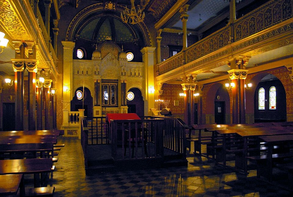 Ilustracja przedstawia wnętrze synagogi. Frontalnie łukowata wnęka z Torą przechowywaną w „świętej skrzyni”, wbudowaną we wschodnią ścianę synagogi. Wnęka przesłonięta jest zdobioną zasłoną zwaną parochet. Nad wnęką wisi lampka – Wieczne Światło. Obok stoi menora, siedmioramienny świecznik. Centralnym miejscem jest podium, podwyższenie, na którym znajduje się pulpit służący do czytania Tory. W dwóch rzędach krzesła z pulpitami dla modlących się i studiujących Torę.