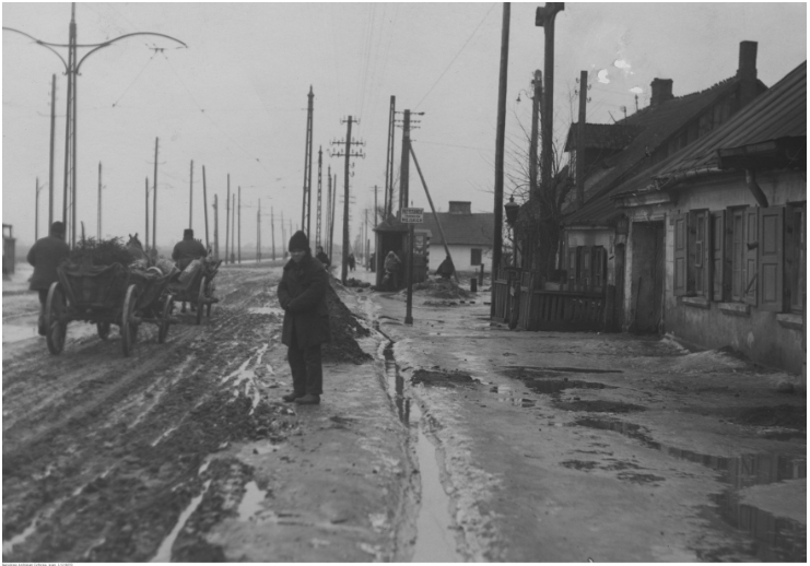 Zdjęcie przedstawia ulicę Grójecką. Ulica to klepisko, pełne błota. Ulicą jadą wozy zaprzężone w konie. Po prawej stronie ulicy znajdują się stare, drewniane domy. Przed domami nie ma chodnika.