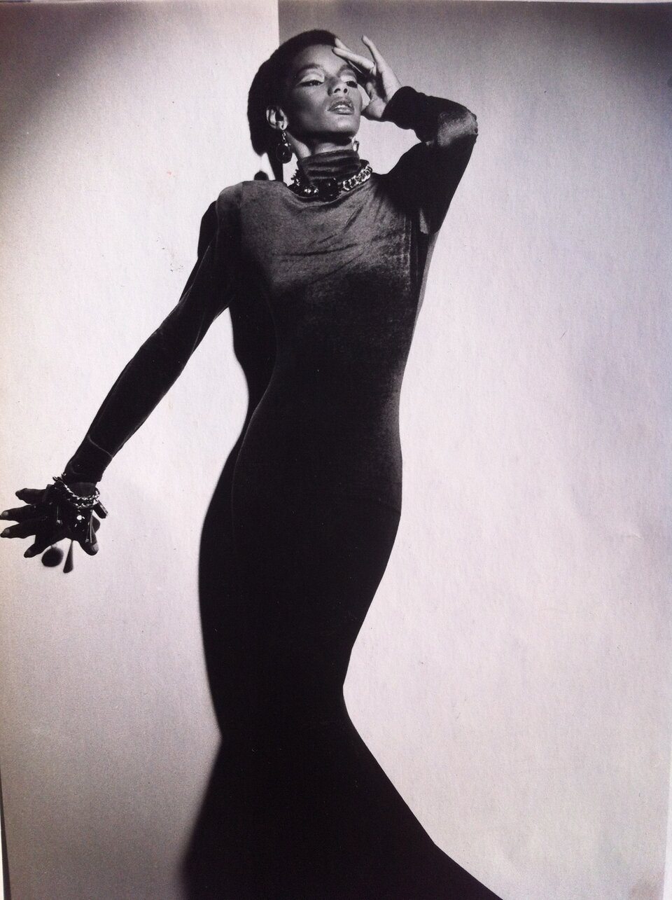 Czarno‑biała fotografia przedstawia czarnoskórą kobietę, pozującą na tle jasnej ściany. Ma ona na sobie długą suknię, rozkloszowaną na dole, z długimi rękawami oraz z golfem. Na jej szyi oraz prawym nadgarstku widoczna jest biżuteria składająca się z grubych łańcuchów oraz ozdobnych zawieszek. Wiszące kolczyki są w podobnym stylu. Kobieta ma uniesioną lekko głowę, patrzy w lewo. Jej powieki pomalowane są jasnymi cieniami, kontrastując z ciemnymi kreskami na górnej linii rzęs. Jej włosy są krótkie. Prawa dłoń kobiety oparta jest o ścianę za nią, a lewa, uniesiona, spoczywa na jej twarzy.