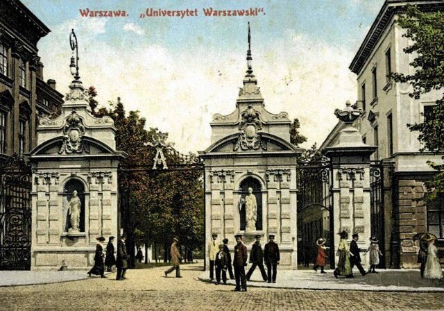 Zdjęcie przedstawia bramę wejściową do budynku Uniwersytetu Warszawskiego. Kute żelazne bramy umocowane są pomiędzy prostokątnymi, szerokimi filarami z rzeźbami umieszczonymi w półkolistych wgłębieniach. Powyżej umieszczone są latarnie wiszące na wygiętych spiralnie prętach. Na jednej z bram duża litera A. Przed wejściem spacerują ludzie. W tle widać budynki i drzewa. 