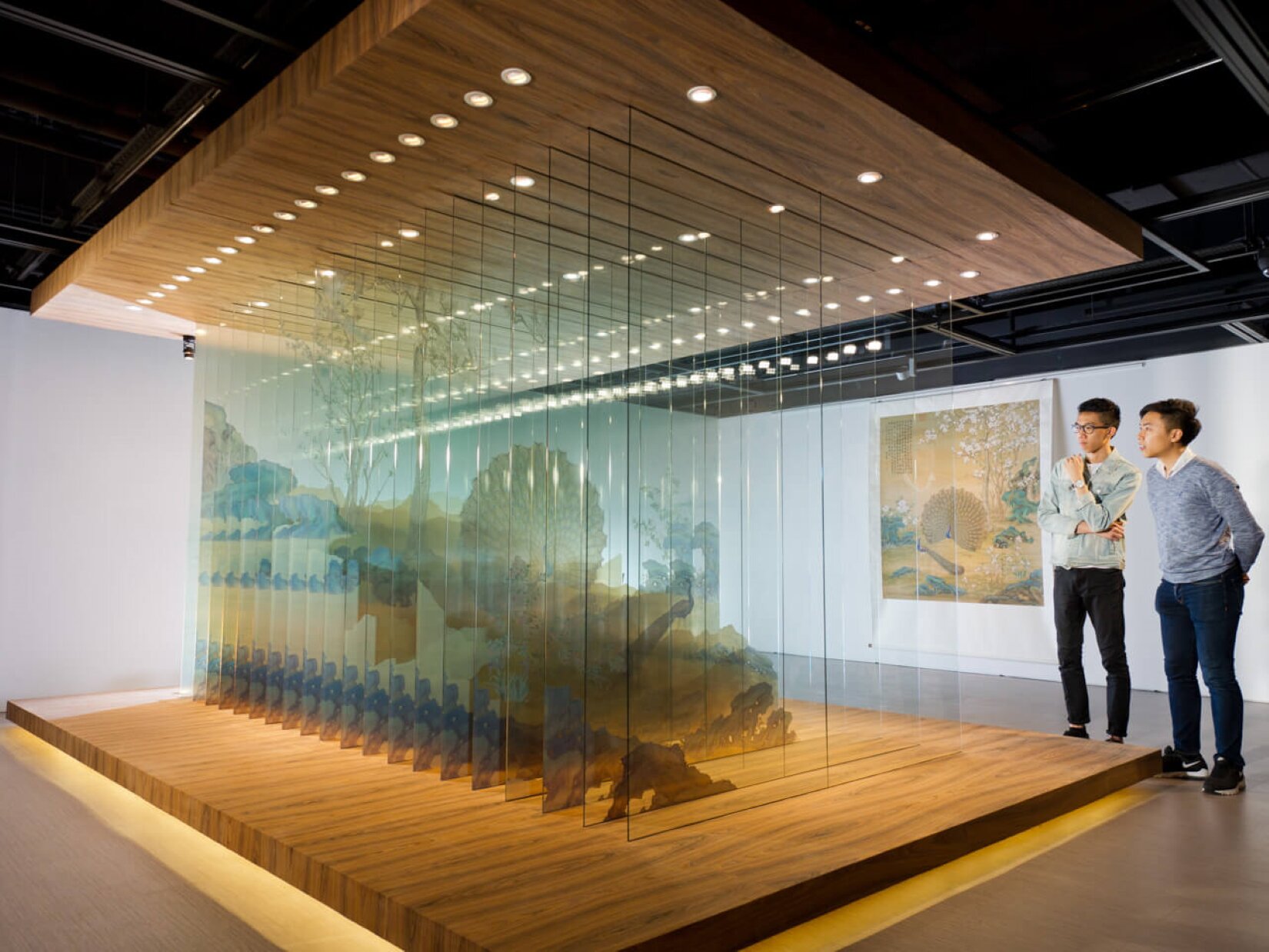 Ukazana została fotografia, na której znajduje się wystawa wykonana z szyb. Na każdej z nich znajduje się jej osobny fragment. Zamocowane są pomiędzy drewnianymi podestami. Od góry oświetlone są punktowymi światłami. Wystawę ogląda dwóch chłopców o ciemnych włosach. Ubrani są w jasnoniebieskie swetry oraz ciemne spodnie. Jeden z nich nosi okulary. Ściany pomieszczenia są białe, a podłoga wykonana jest z drewna. Na tylnej ścianie zawieszony jest obraz, który przedstawia efekt, który powinna stworzyć wystawa na szybach.