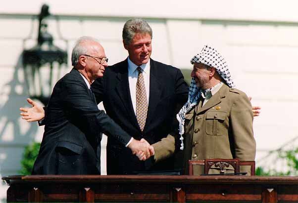 Na zdjęciu znajduje się Icchak Rabin i Jasir Arafat, którzy ściskają sobie dłonie. Za nimi stoi Bill Clinton, który ich obejmuje. Clinton i Rabin są ubrani w ciemne garnitury, a Arafat w jasna marynarkę i białą chustę. Przed nimi stoi drewniane biurko. 