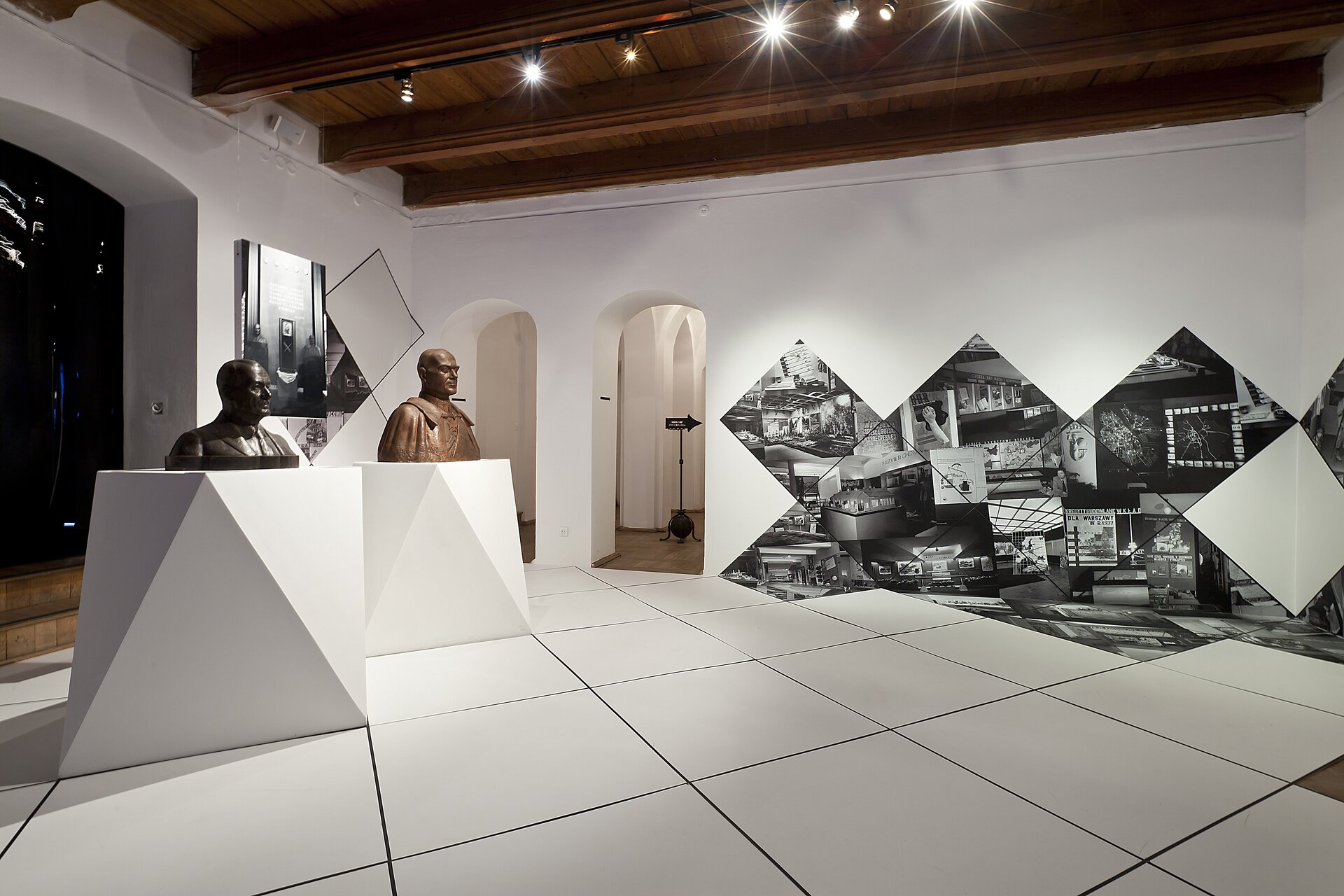 Na zdjęciu znajduje się pomieszczenie o brązowym, drewnianym suficie, białych ścianach i podłodze w białe, duże kafle. Po lewej stronie znajdują się 2 geometryczne, białe postumenty. Na pierwszym jest popiersie mężczyzny w kolorze ciemnoszarym, a na drugim popiersie mężczyzny w kolorze brązowym. Na ścianie za rzeźbami są biało‑czarne ilustracje. Po prawej stronie znajdują się biało‑czarne ilustracje na płytach w kształcie czworoboku. W tle są 2 wąskie przejścia do kolejnego pomieszczenia. Na suficie są małe, czarne lampki. 