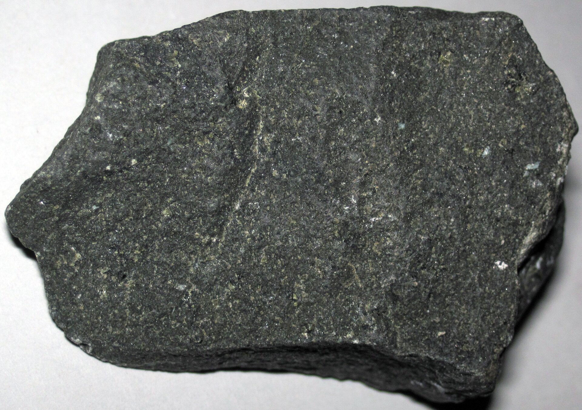 Zdjęcie przedstawiające kawałek bazaltu. Skała ma ciemnoszarą barwę i drobną strukturę krystaliczną. Miejscami na jej powierzchni występują rzadko rozmieszczone białe fragmenty.