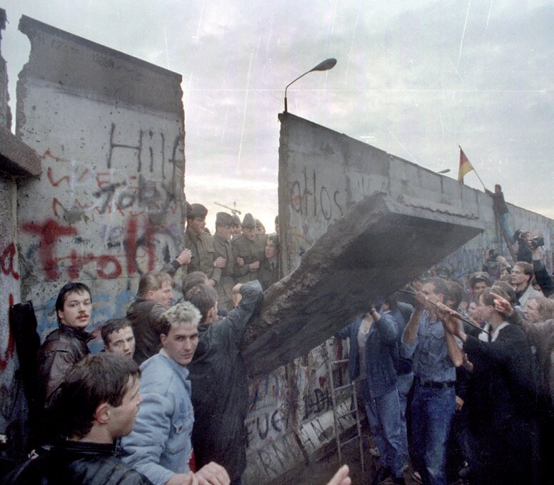 Zdjęcie przedstawia betonowy mur, pod którym zgromadzona jest grupa ludzi. Niszczą fragment muru. Zza zniszczonego fragmentu, po drugiej stronie muru, widoczna jest grupa żołnierzy w mundurach.