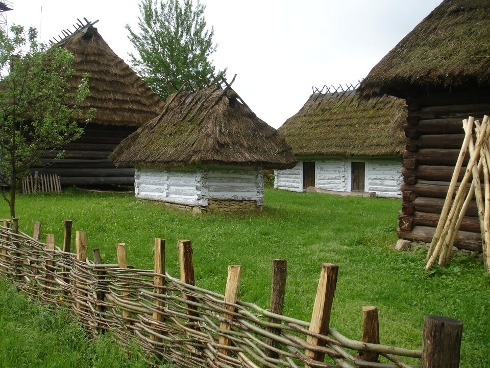 Zdjęcie przedstawia chałupy. Na pierwszym planie znajduje się pleciony drewniany płot. Dalej jest trawnik, na którym stoją chałupy. Są one zbudowane z poziomo ułożonych na sobie drewnianych desek. Niektóre domy mają je pomalowane na białe a inne naturalne. Dach posiada słomę.