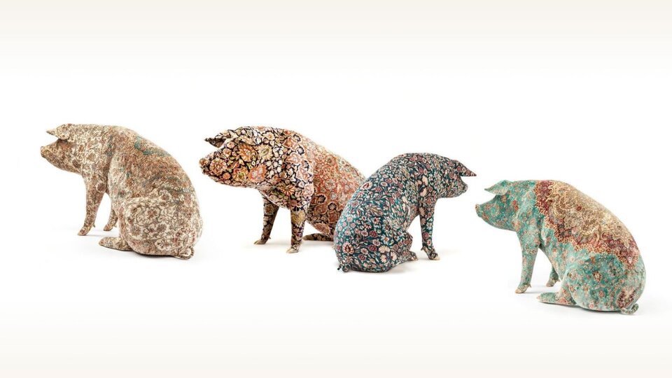 Fotografia przedstawia figurki czterech świń. Zwierzęta pokazano w pozycji siedzącej, od tyłu, każde jest widoczne z innej perspektywy. Każda świnia jest w innym kolorze. Od lewej: Pierwsza świnia ma tatuaż w kolorze beżu z białymi wzorami i kwiatami, druga świnia również została ozdobiona kwiecistymi ornamentami w kolorze ciemnoniebieskim oraz pomarańczowym i beżowym. Trzecia świnia ma tatuaż o ciemnoniebieskim tle, na którym umieszczono czerwone, beżowe i żółte kwiaty. Czwarta świnia ma tatuaż z błękitnym tłem, na grzbiecie - czerwonym i brązowym, na którym umieszczono białe i czerwone kwiaty.  Świnie zostały ustawione jedna obok drugiej. Tatuaże na ich skórach przypominają wzory znajdujące się na dywanach. 