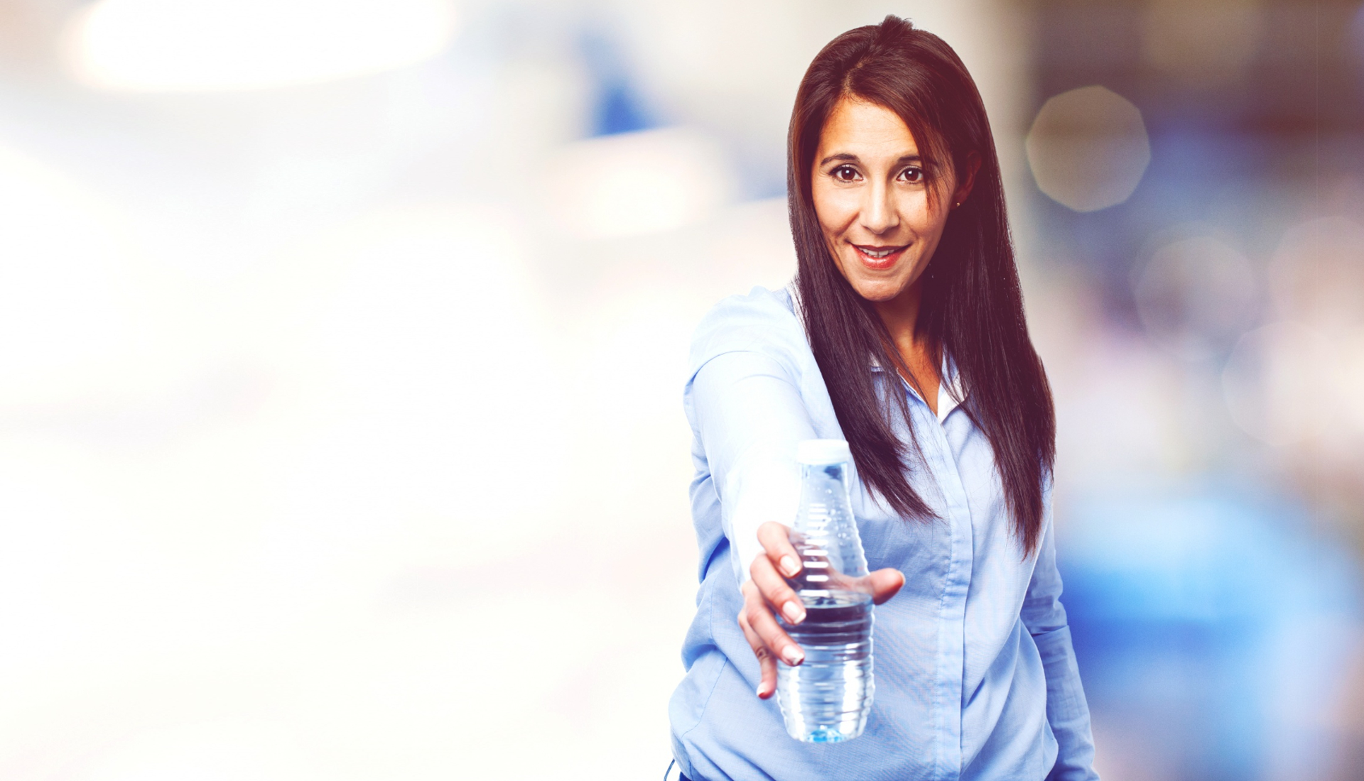 Zdjęcie przedstawia kobietę, która wysuwa prawą dłoń w kierunku obiektywu. Trzyma w niej butelkę wody. 