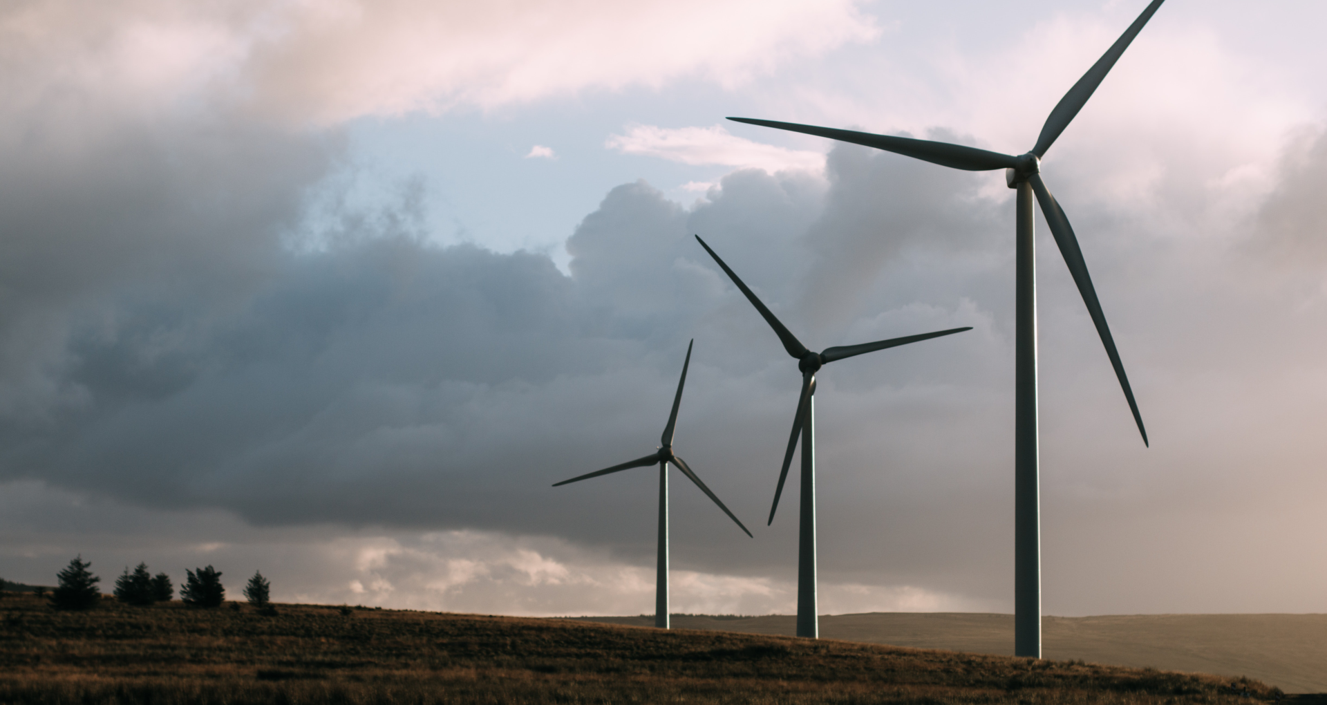 Zdjęcie przedstawia trzy turbiny wiatrowe. Każda turbina składa się z wąskiego pionowego cokołu, na którym widać trzy śmigła ułożone równomiernie w polu koła. W tle zachmurzone niebo.
