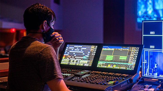 Na zdjęciu znajduje się siedzący na krześle mężczyzna z czarną maseczką na twarzy. Siedzi on przed konsolą do obróbki dźwięku składającej się z różnych suwaków, pokręteł i dwóch ekranach wyświetlających słupki w aplikacji do edycji dźwięku.