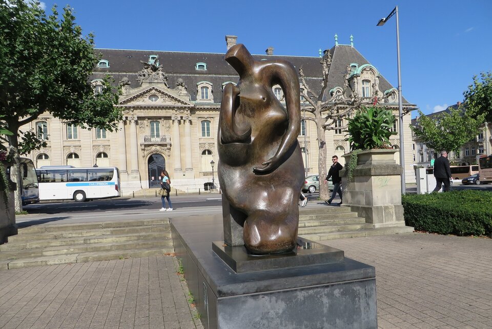 Zdjęcie przedstawia rzeźbę wykonaną w brązie. Rzeźba o bryle zwartej, przedstawia matkę i dziecko. W tle znajduje się budynek oraz wyłożony kostką plac. Po obu stronach znajdują się krzewy oraz drzewa, częściowo ucięte przez kadr fotograficzny.