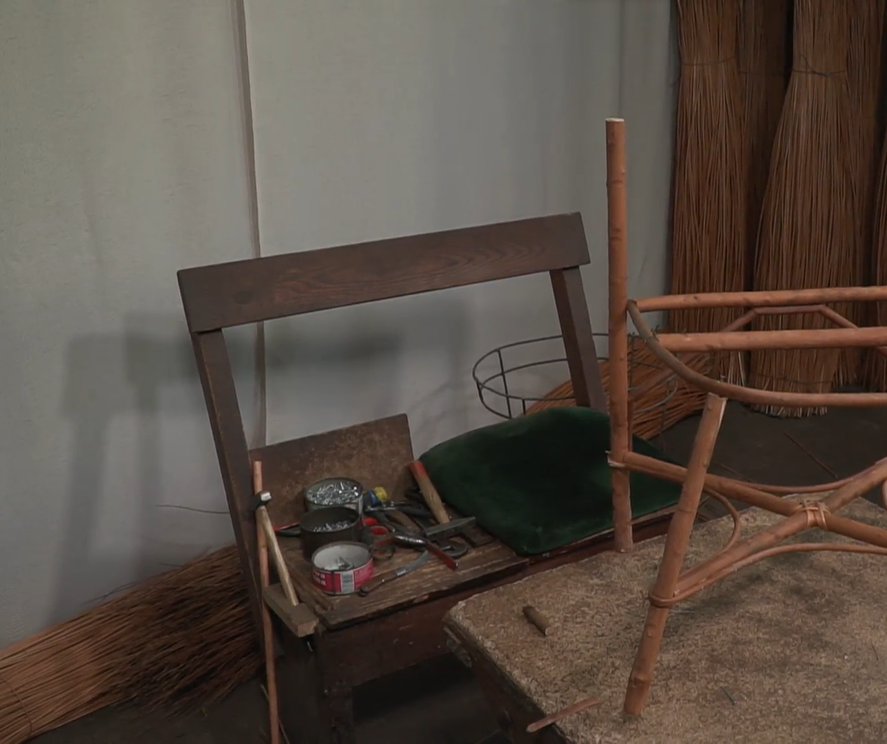 Zdjęcie przedstawia ławkę z narzędziownikiem, która stoi przy stole. Na ławce leżą narzędzia plecionkarskie oraz poduszka. 