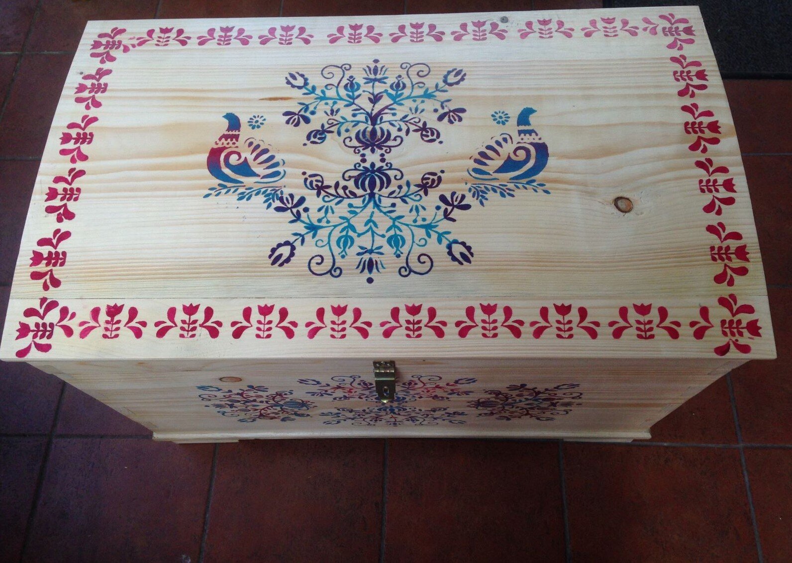 Zdjęcie wykonane zostało z góry i przedstawia kufer z drewna, z metalowym zamknięciem, malowany w ludowe ornamenty. Symetryczny wzór na wieku kufra składa się z gałązek, listków i pąków kwiatów, po bokach dwa gołąbki. Całość obiega szlak z powtarzającego się motywu kwiatka z listkami. Przednia ścianka pod zamkiem jest również malowana, ale wzór jest niewyraźny.